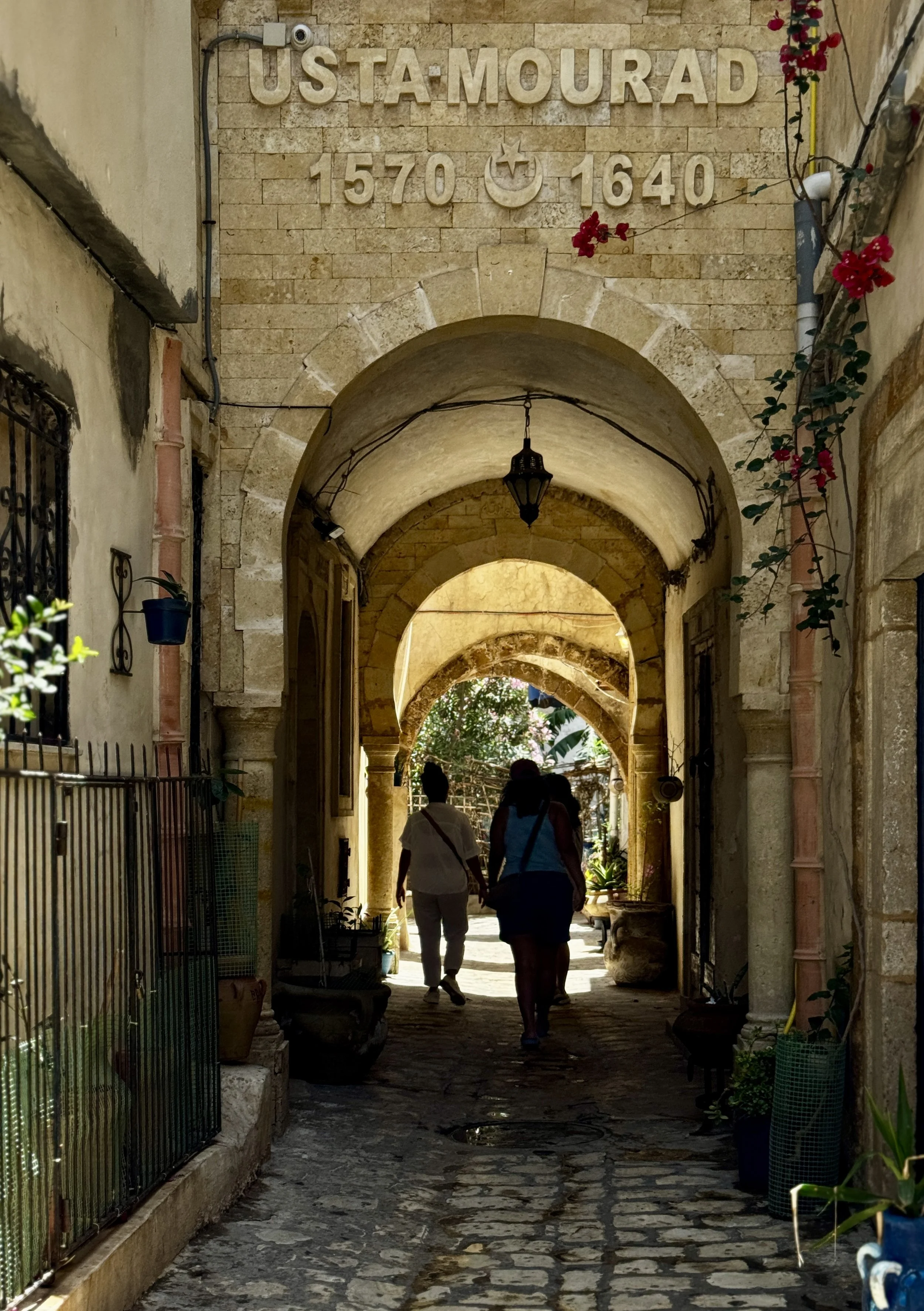 People walking through a stone archway with a sign that reads "Ustá-Mourad 1570 + 1640" on a beige brick wall. The scene is lit by a hanging lantern, and there are plants and vines along the sides.
