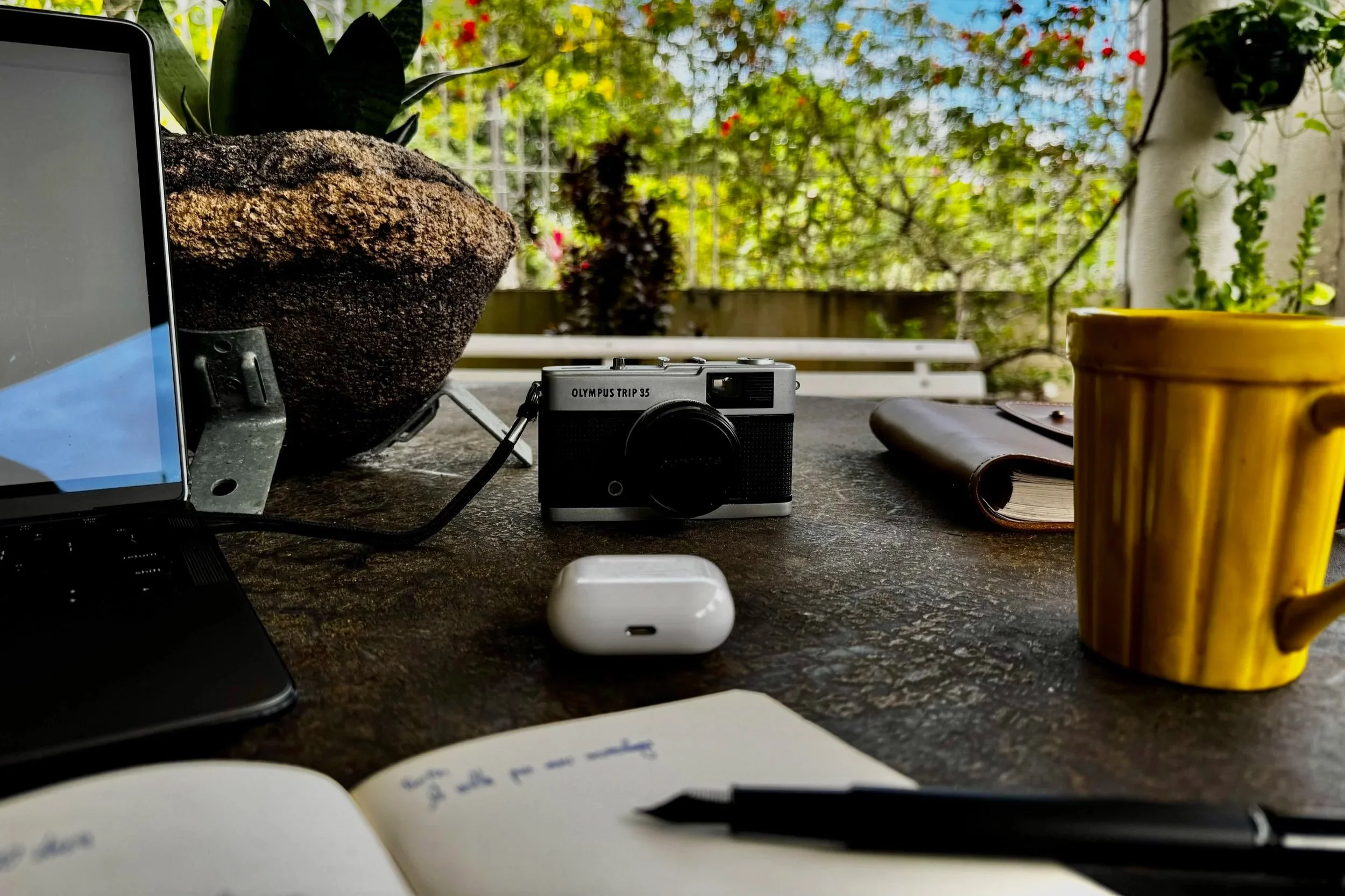 A cluttered outdoor workspace with a camera, laptop, notebook, AirPods, a yellow mug, and a plant pot on a table, with a background of greenery and a fence.
