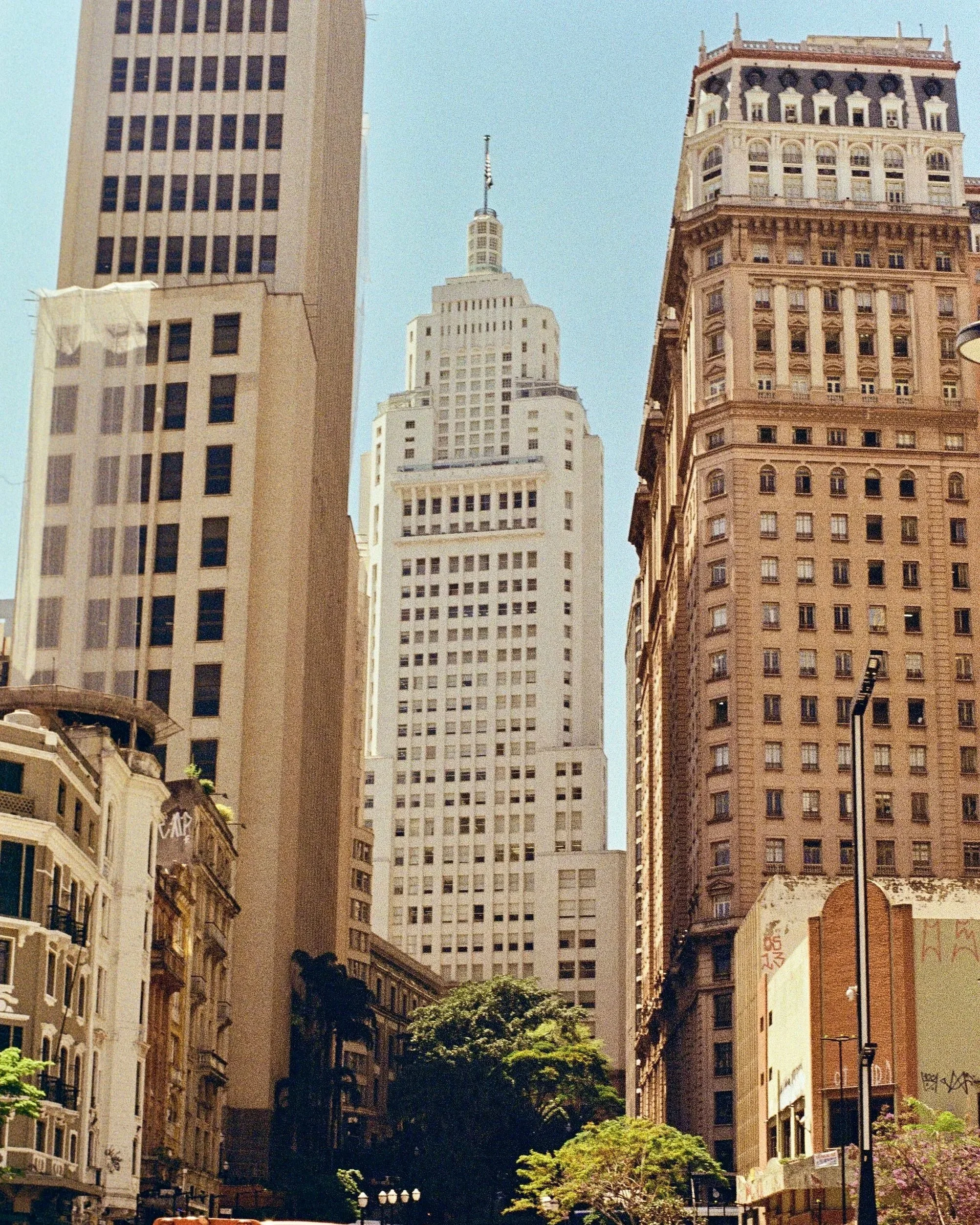 Tall skyscraper in downtown city surrounded by shorter buildings, trees, and streetlights.