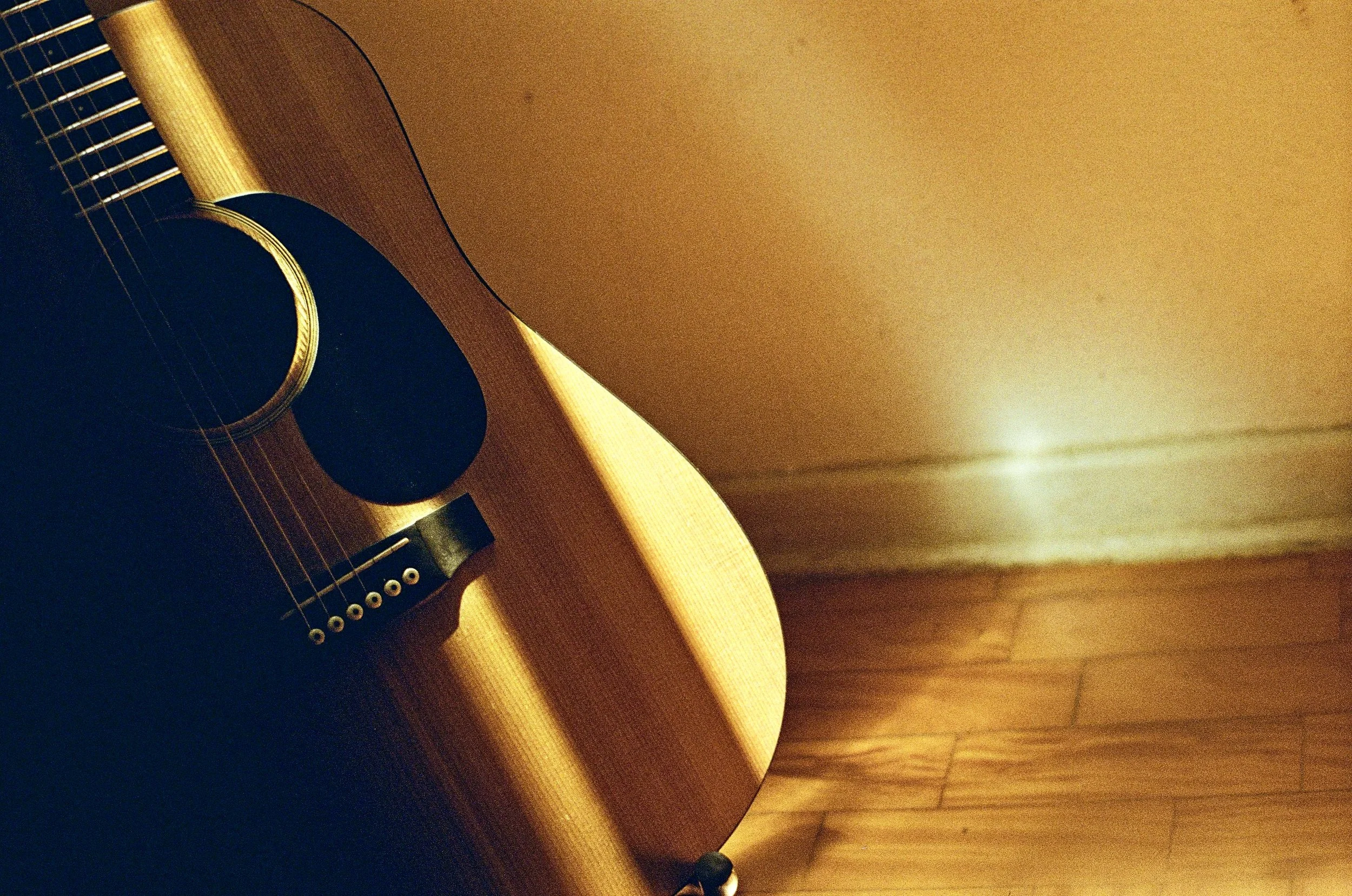Close-up of a wooden acoustic guitar resting against a wall, with a warm ambient light illuminating the scene and a hardwood floor visible for context.