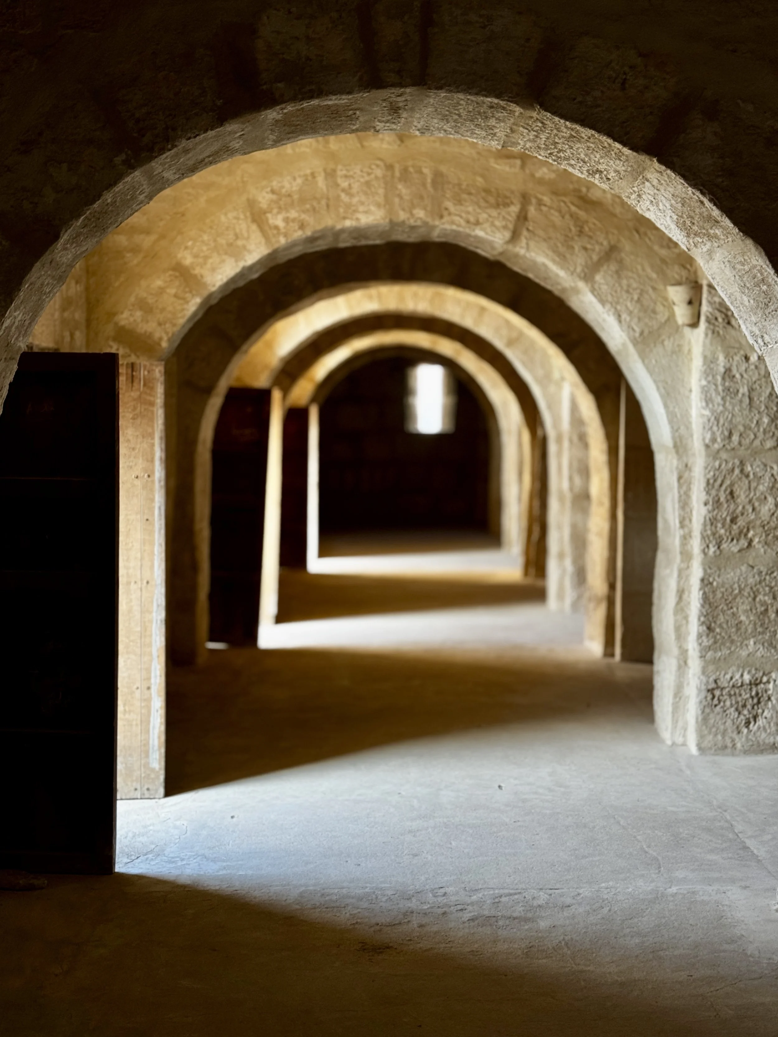 Series of stone arches in a hallway or tunnel, diminishing in size towards the background, with sunlight illuminating the floors and highlighting the texture of the stone.