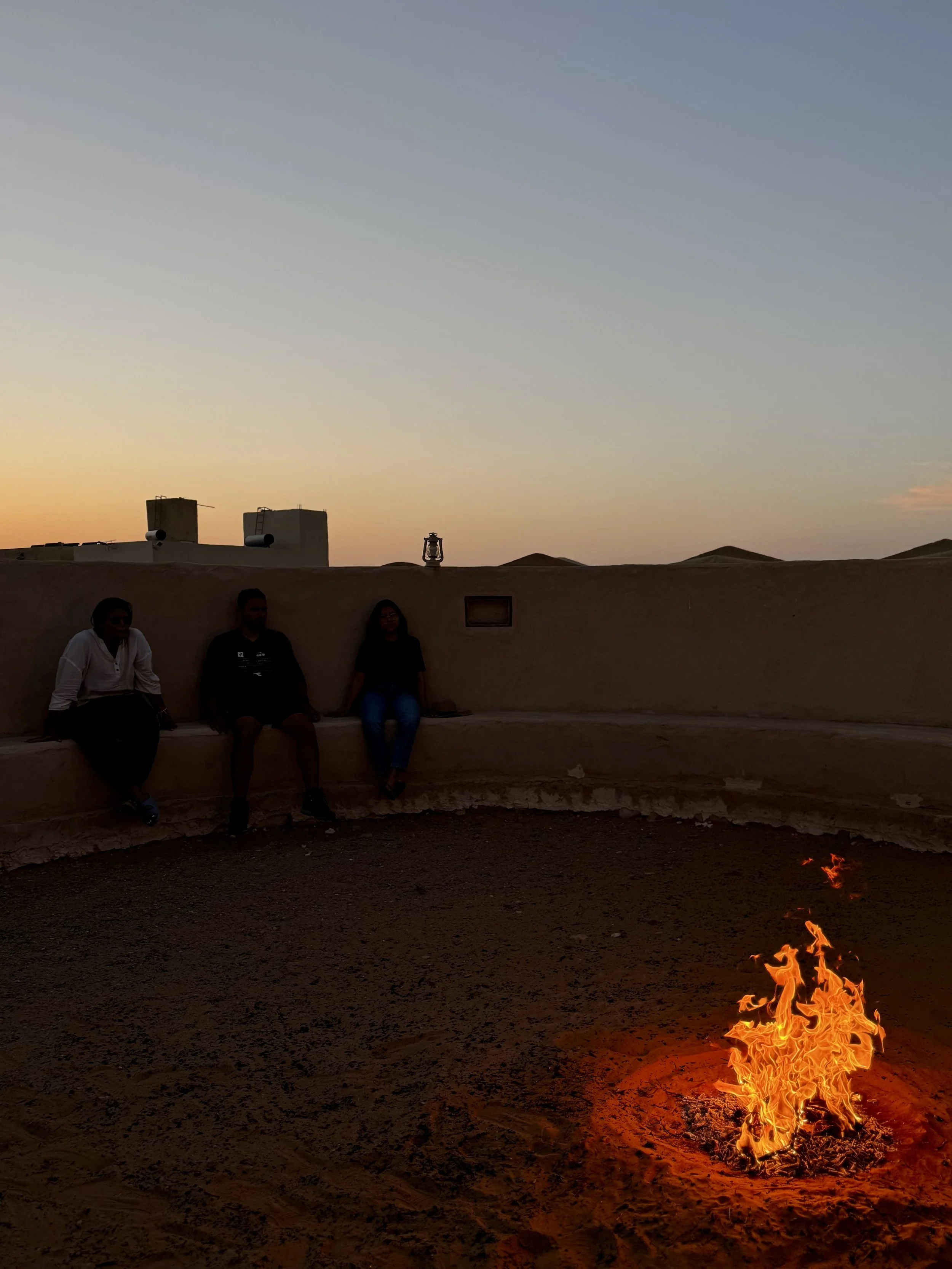 People sitting on a low wall at sunset with a desert sunset sky, structures, a lantern, and a small fire on the ground.