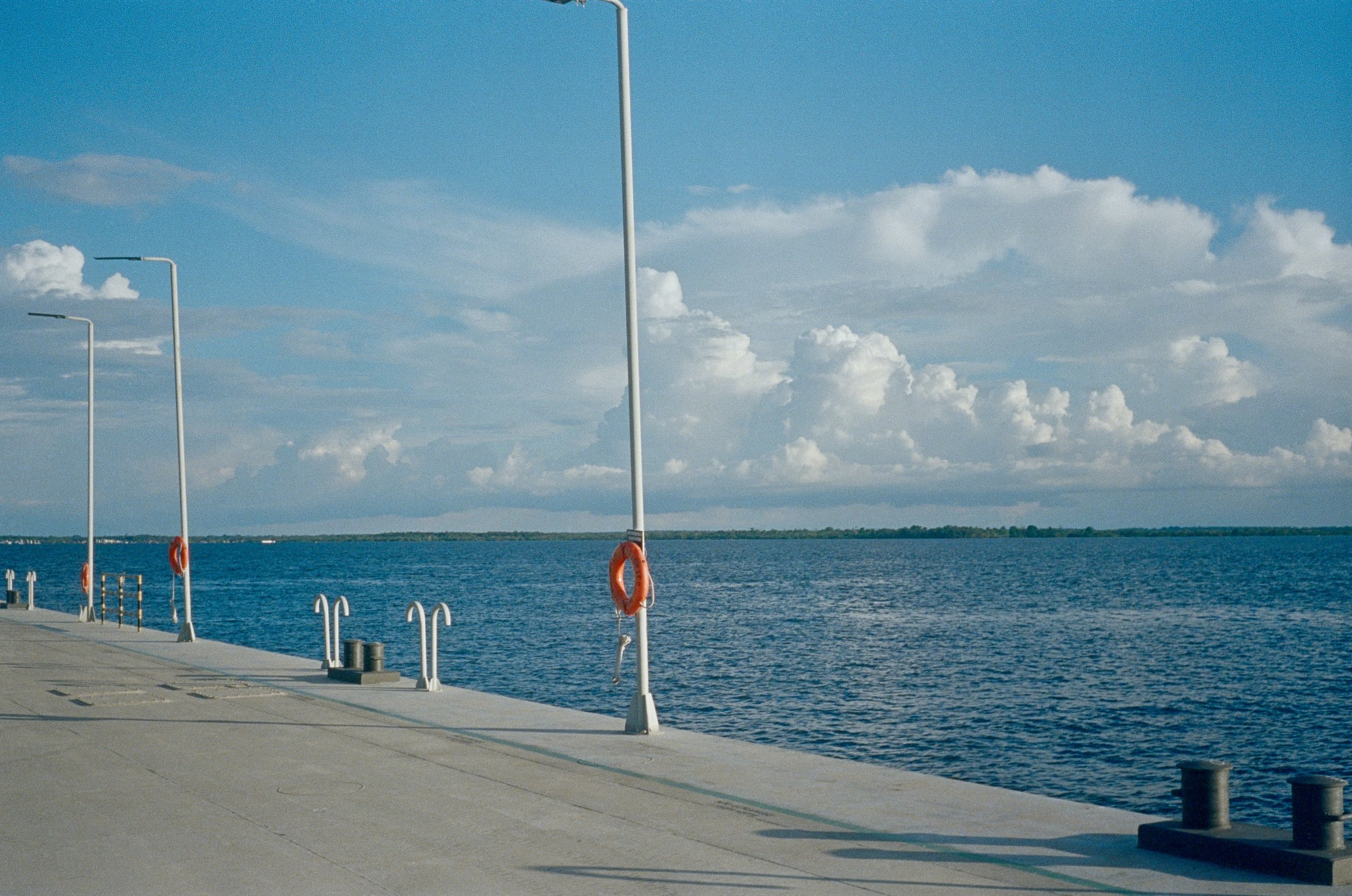 A concrete dock along a large body of water under a partly cloudy sky with several modern light poles and life buoys.
