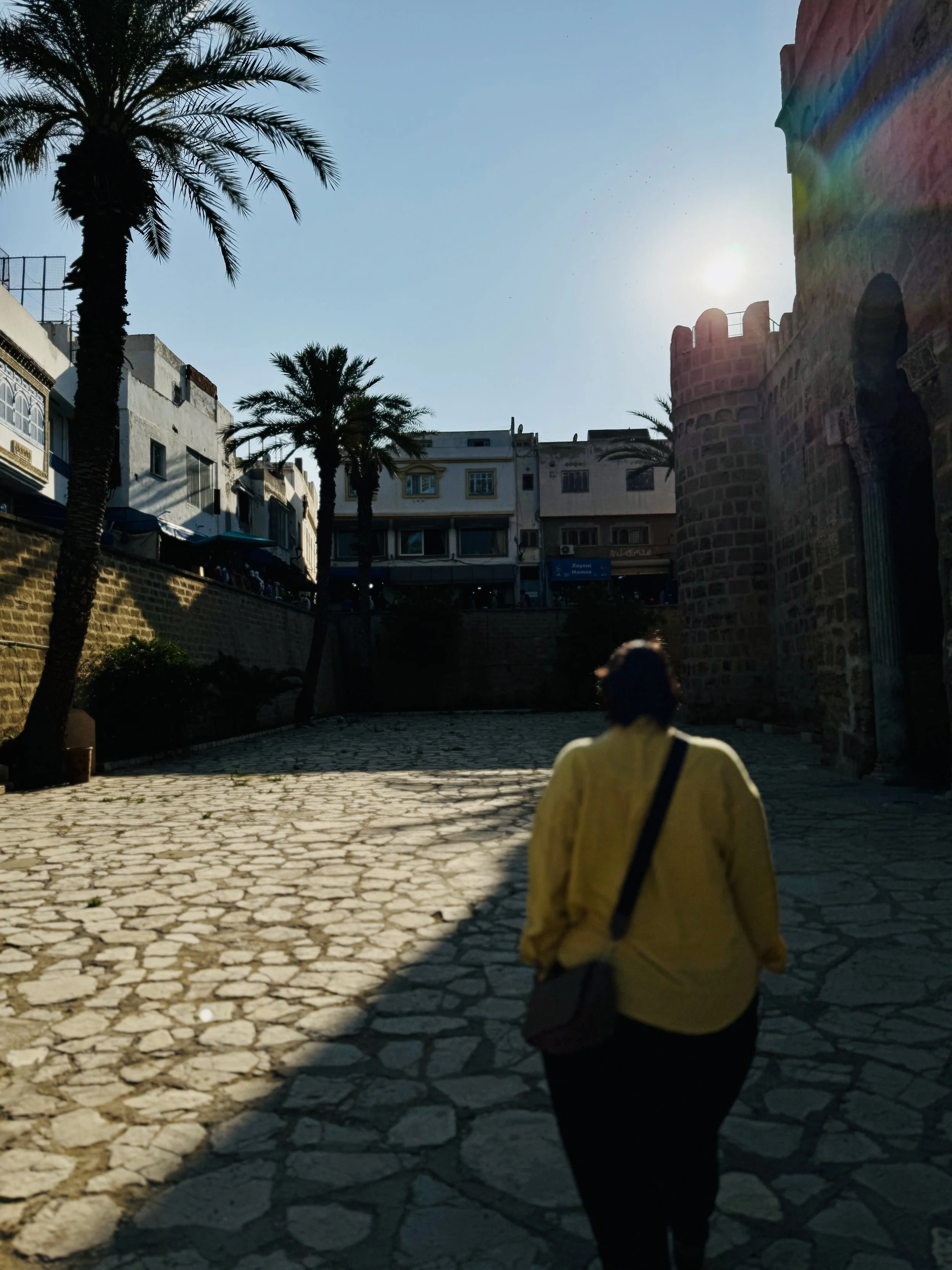 A person walking along a cobblestone street lined with palm trees and historic buildings, with the sun shining brightly in the sky near a castle-like structure.