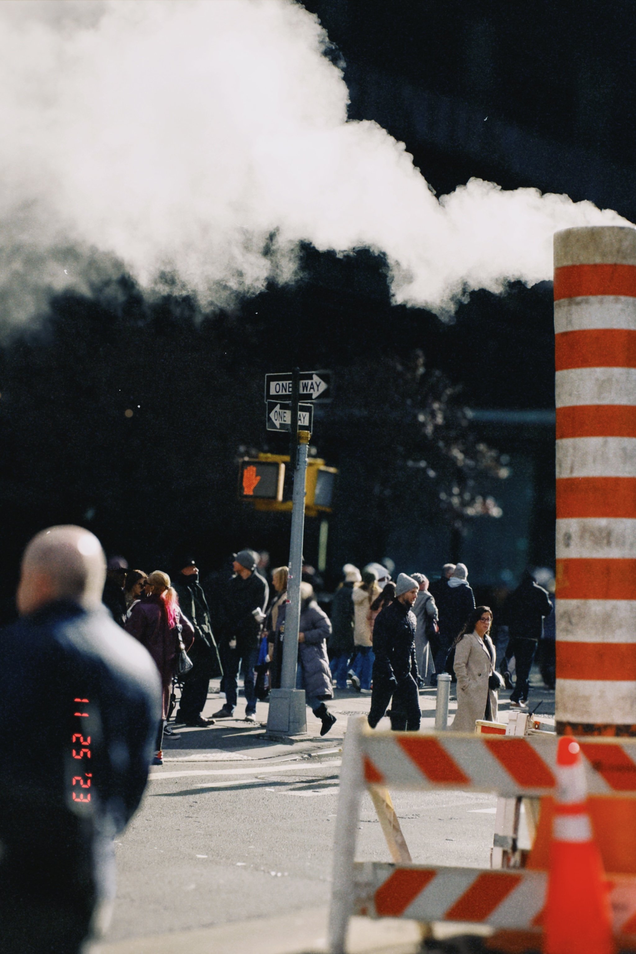 People crossing the street at night with a large plume of smoke or steam coming from a building or factory in the background; traffic signs and construction barriers are visible.