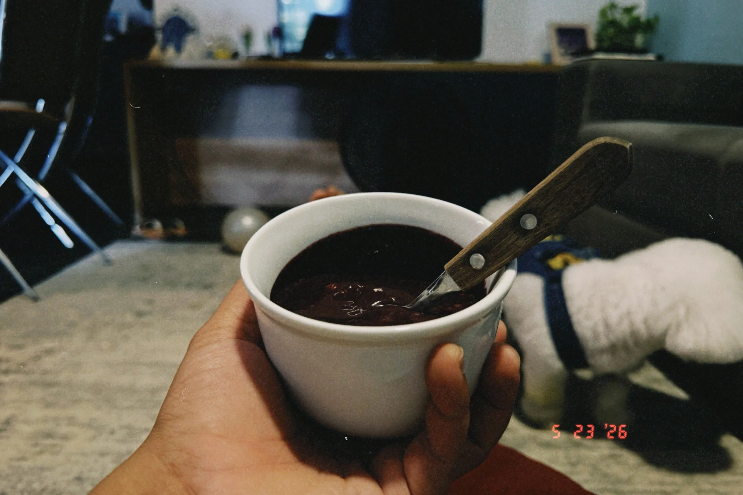 Person holding a white cup filled with dark coffee or hot chocolate, with a spoon resting inside. The background shows chairs, a desk, and some potted plants in a room.