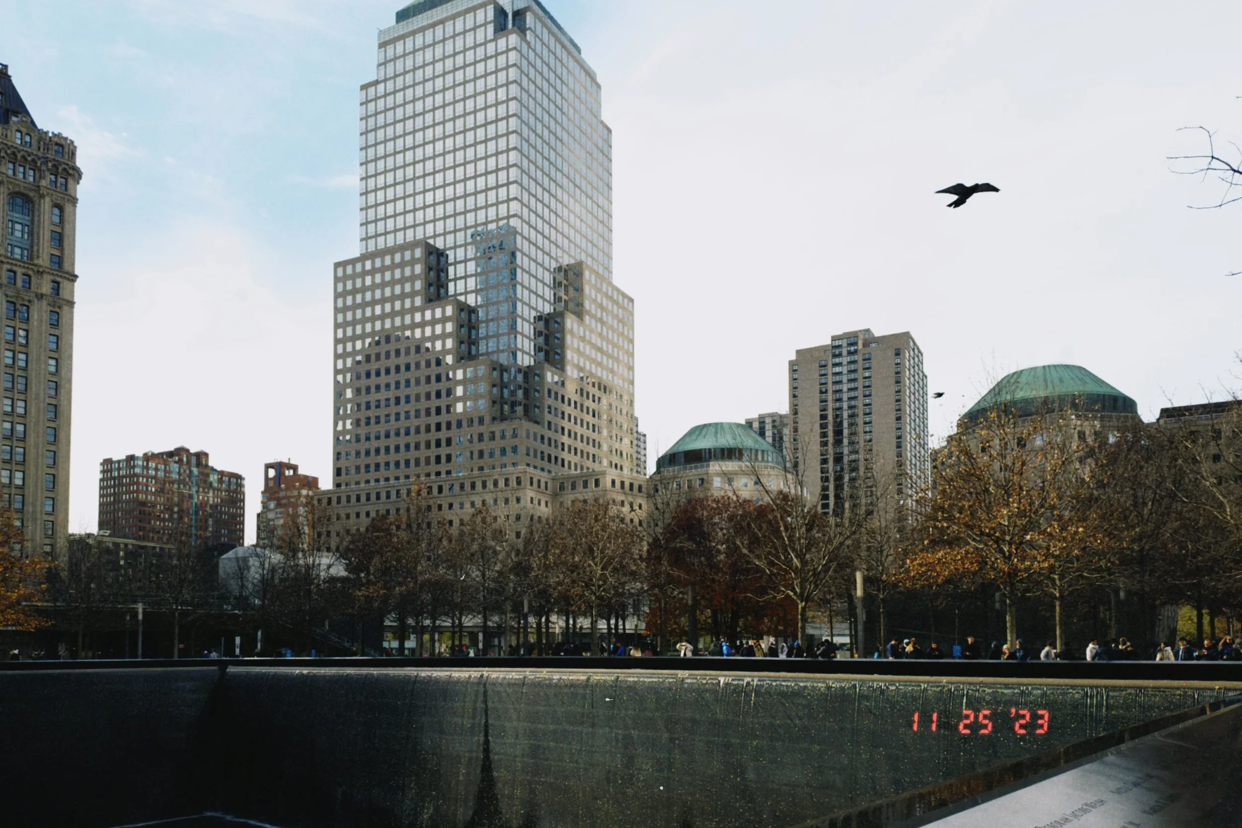 A cityscape with tall buildings, a bird flying in the sky, and a park with trees in the foreground. A digital clock displaying the date and time is at the bottom of the image.