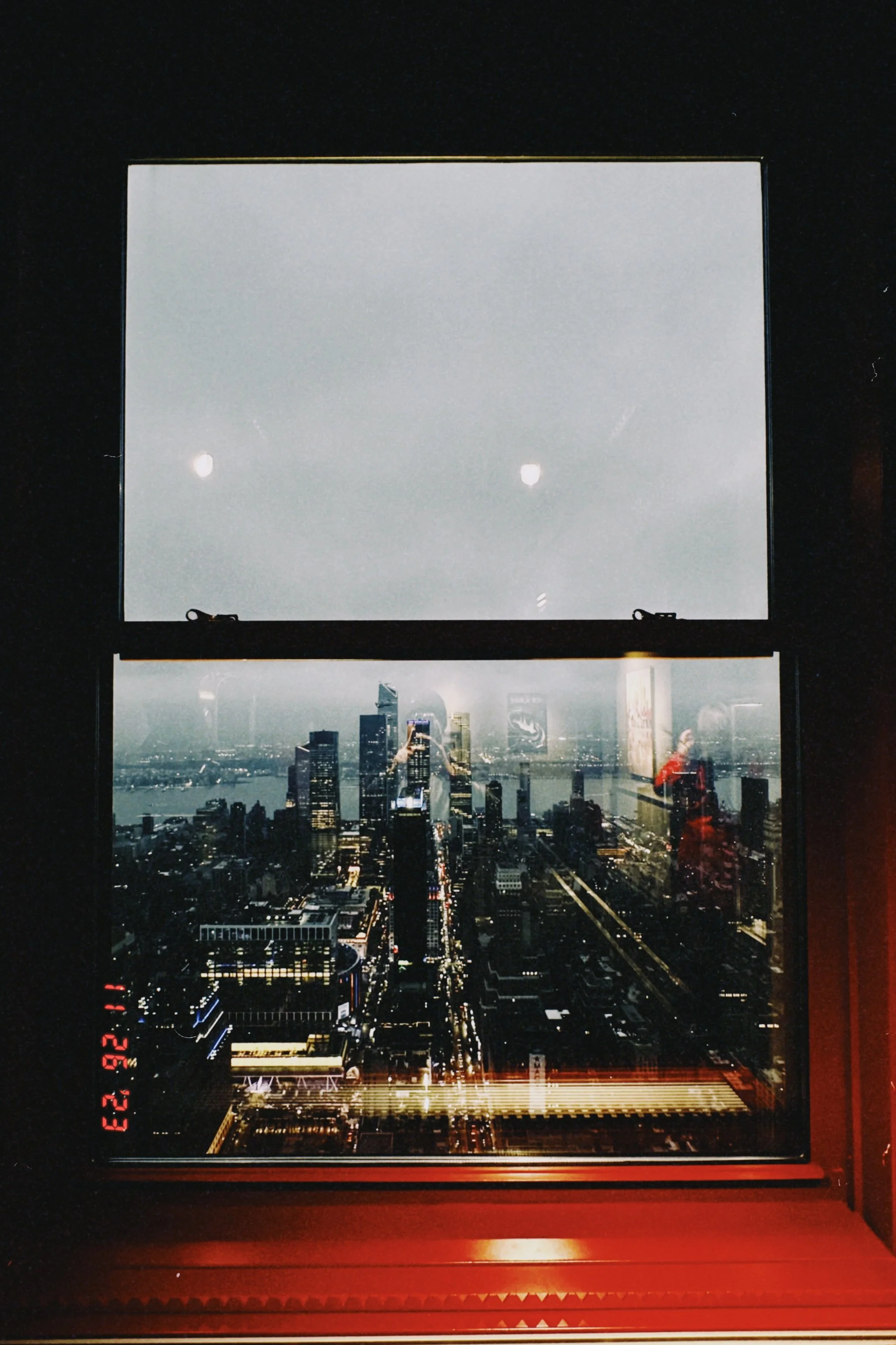 View of a city skyline through a window at night, with illuminated buildings and streets, and a river in the background.