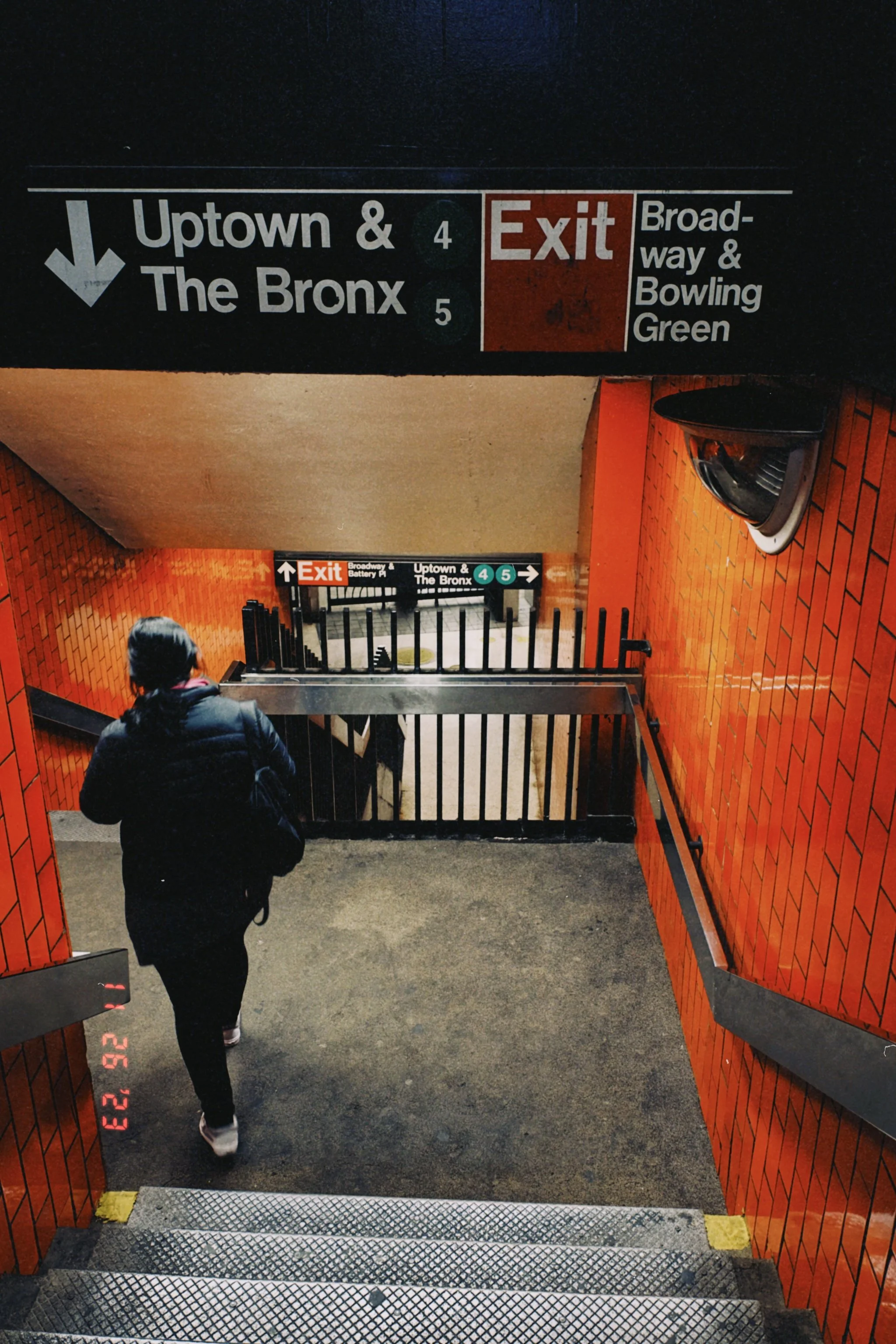 A person with a backpack walking down a staircase at the subway station, with orange brick walls on either side and signs indicating directions to uptown and The Bronx, and an exit to Broadway and Bowling Green.