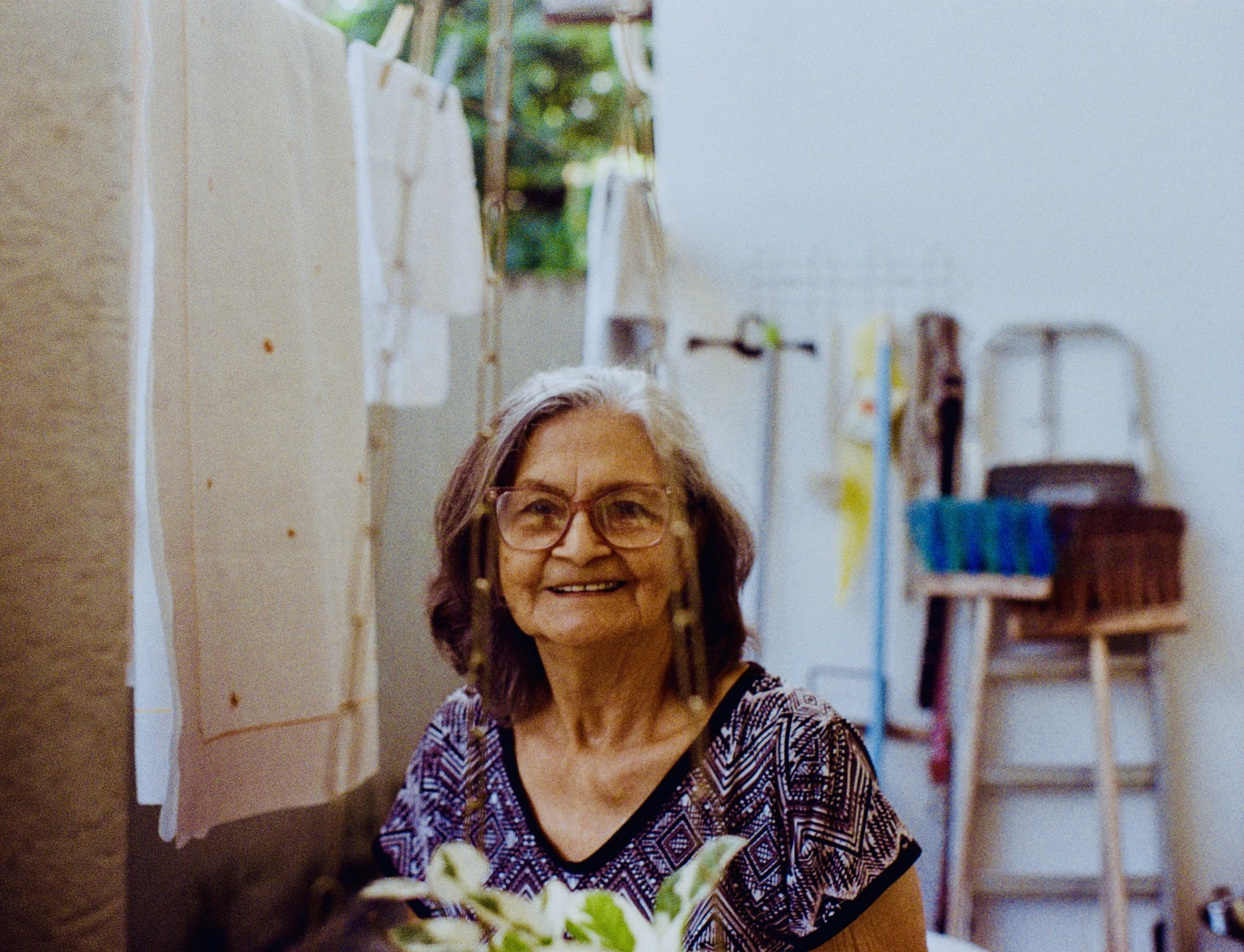 An elderly woman with glasses smiling in a kitchen with shelves and blue wall in the background.
