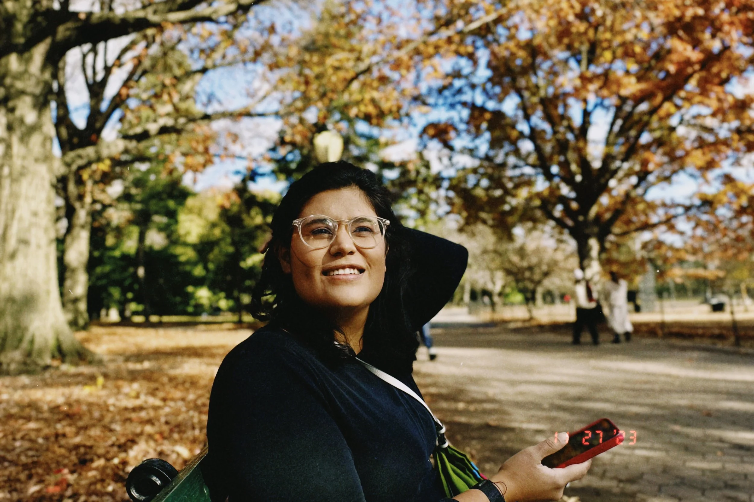 A woman with glasses laughing while sitting outdoors in a park during fall, holding a phone with the temperature display reading 27°C.