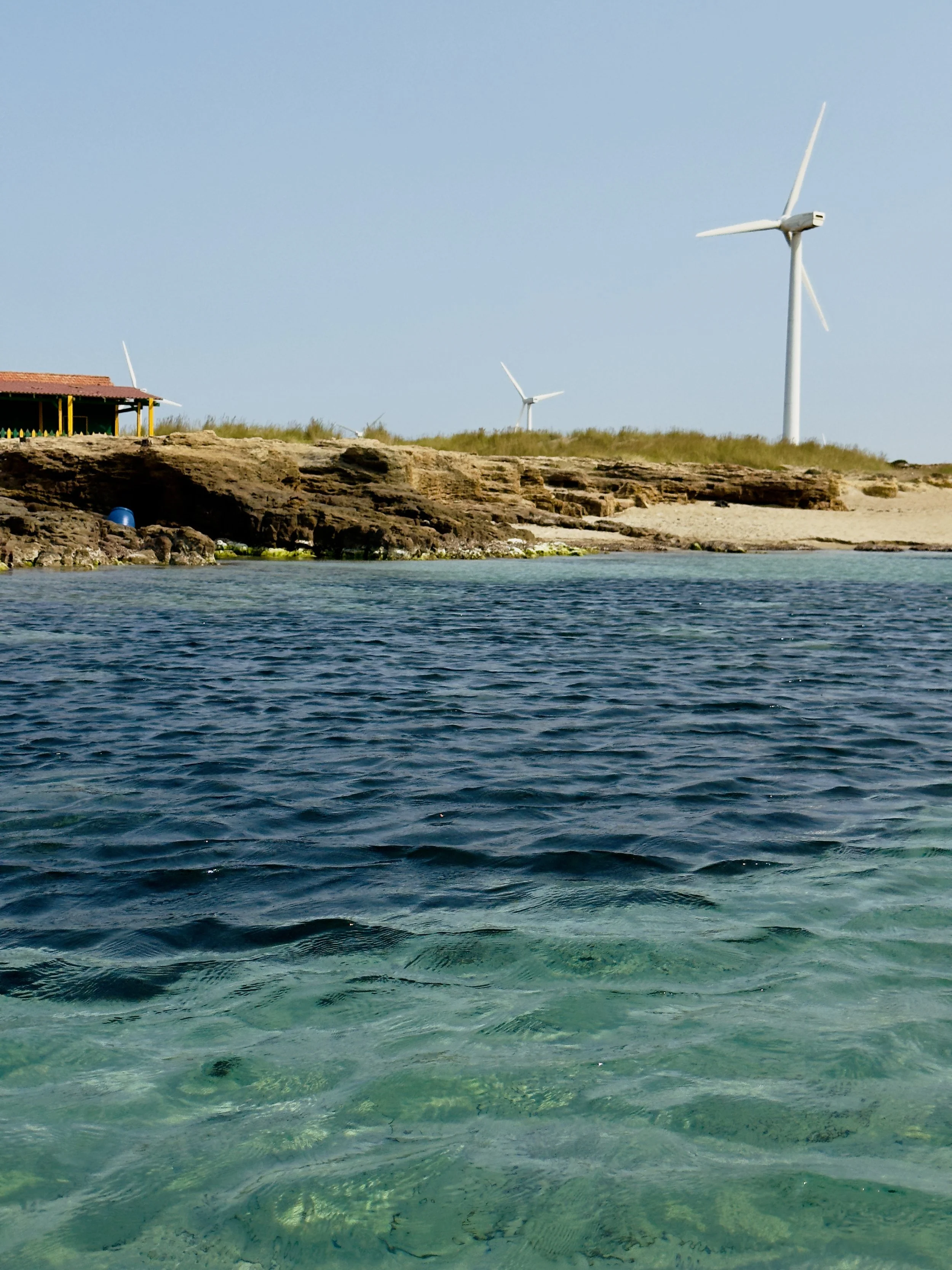 The image shows a body of water with a rocky shoreline. In the background, there are wind turbines and a small structure with a red roof on the land.
