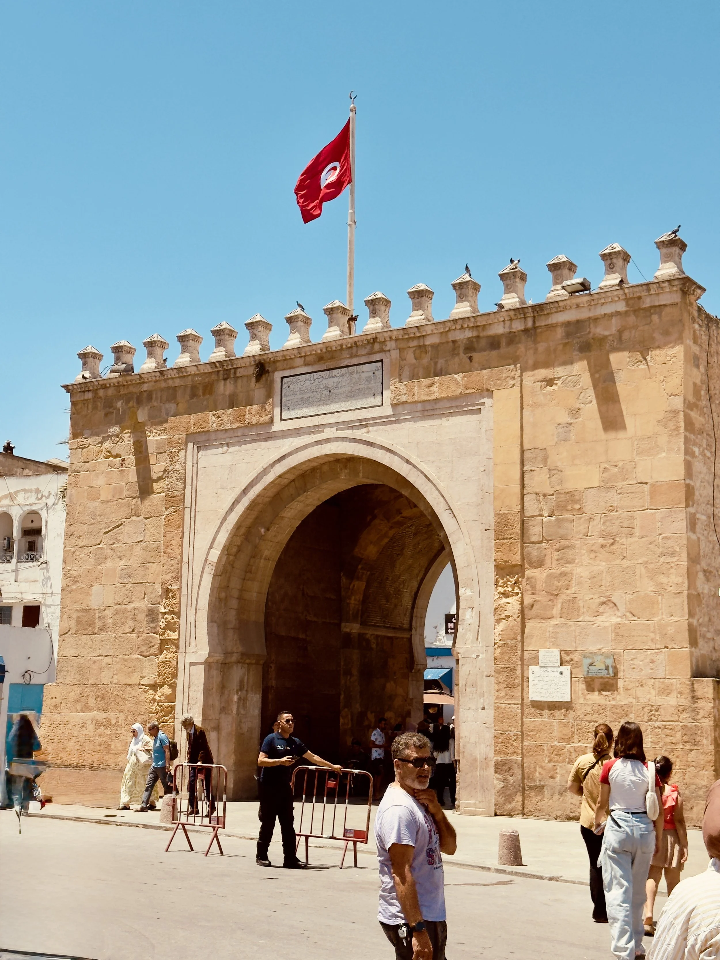 People standing and walking in front of an ancient stone gateway with a Tunisian flag flying on top, against a clear blue sky.