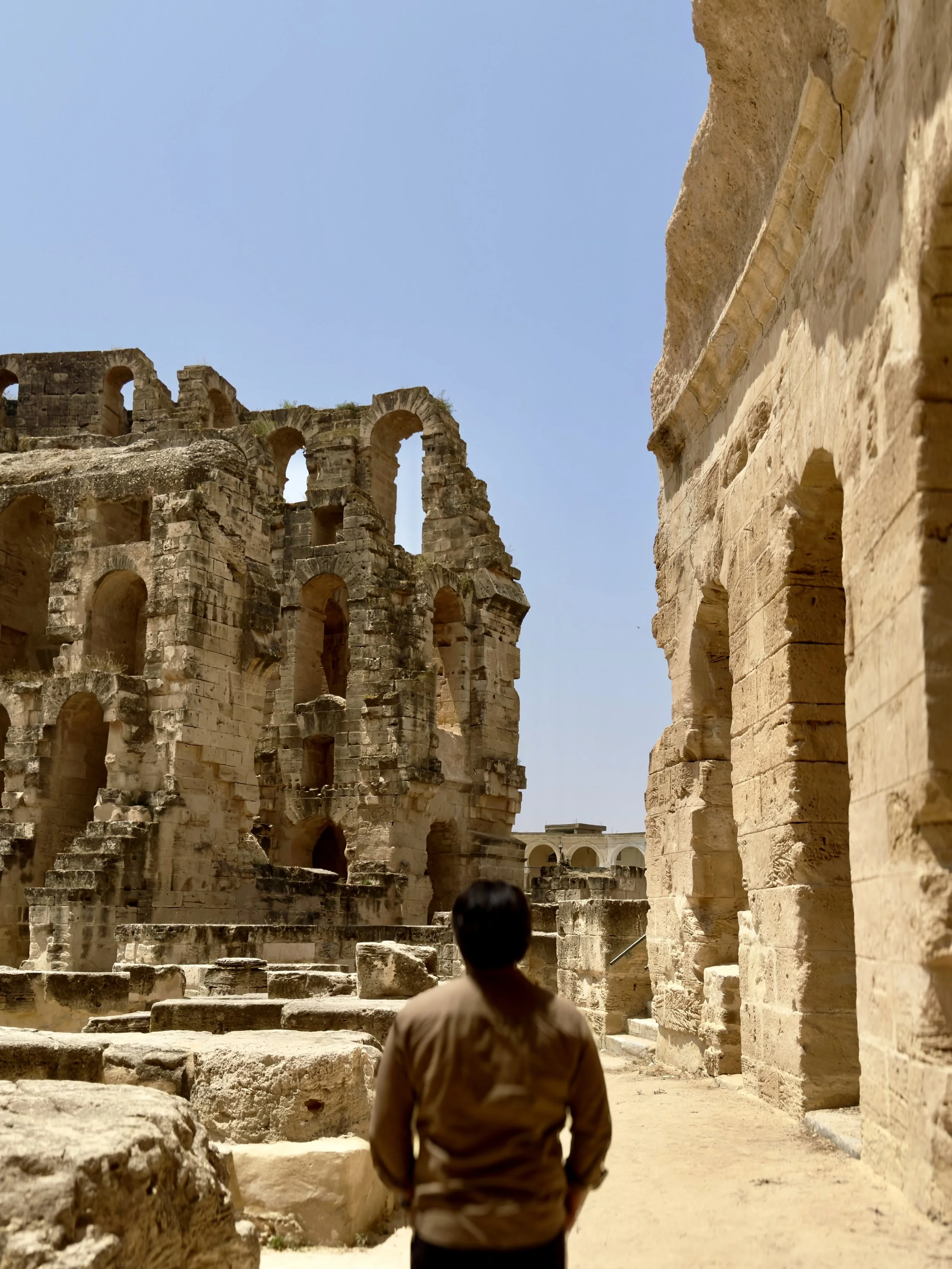 A person with short dark hair and a beige shirt stands in front of ancient stone ruins of the Colosseum in Rome, Italy, with a clear blue sky above.