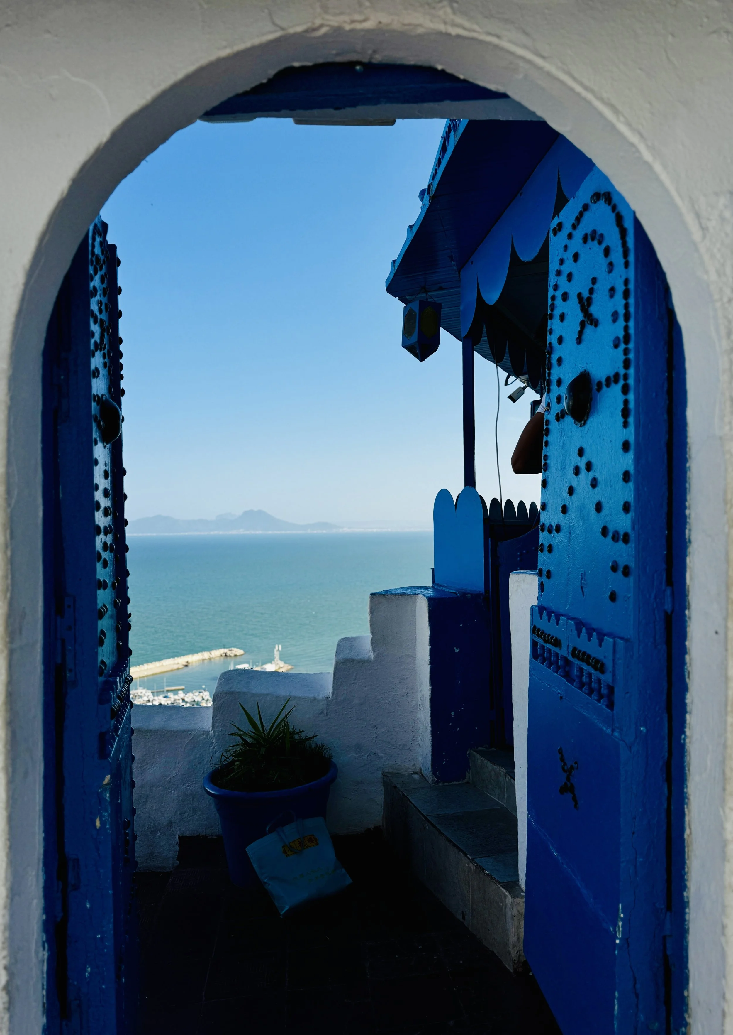 View through a blue and white arched doorway showing a coastal scene with buildings, a harbor, and mountains in the distance under a clear sky.