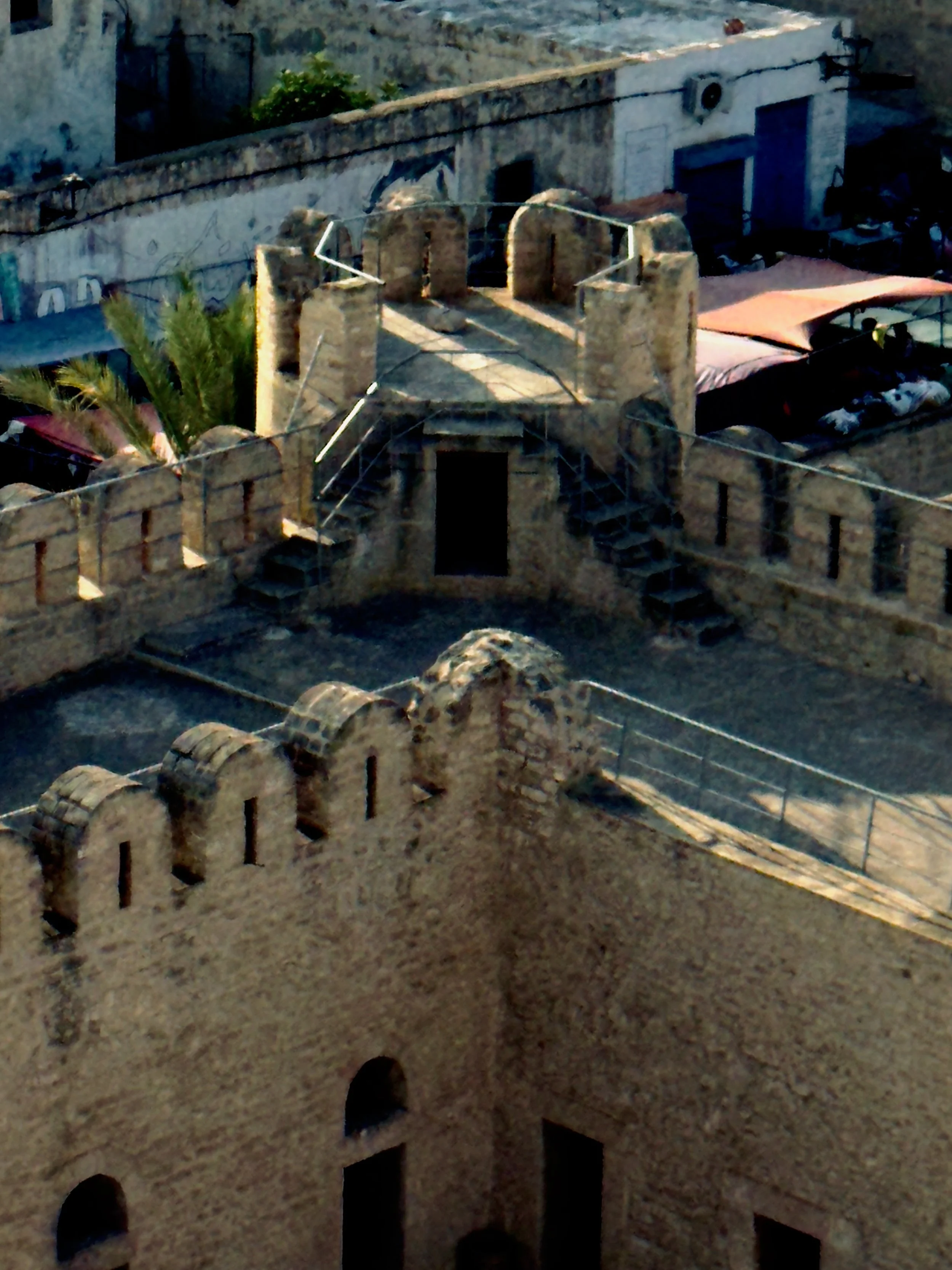 A model or replica of a historic stone fortress with battlements, small towers, and a staircase, situated outdoors against a background of buildings and trees.