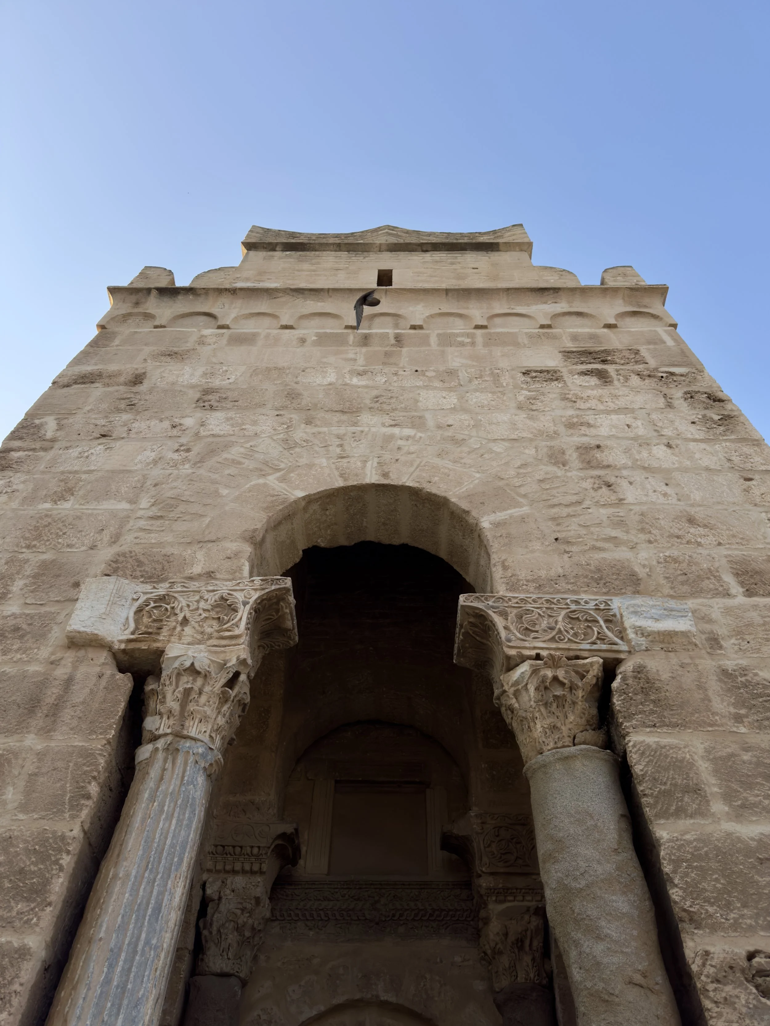 Looking up at an ancient stone structure with arches and carved columns, set against a clear blue sky.