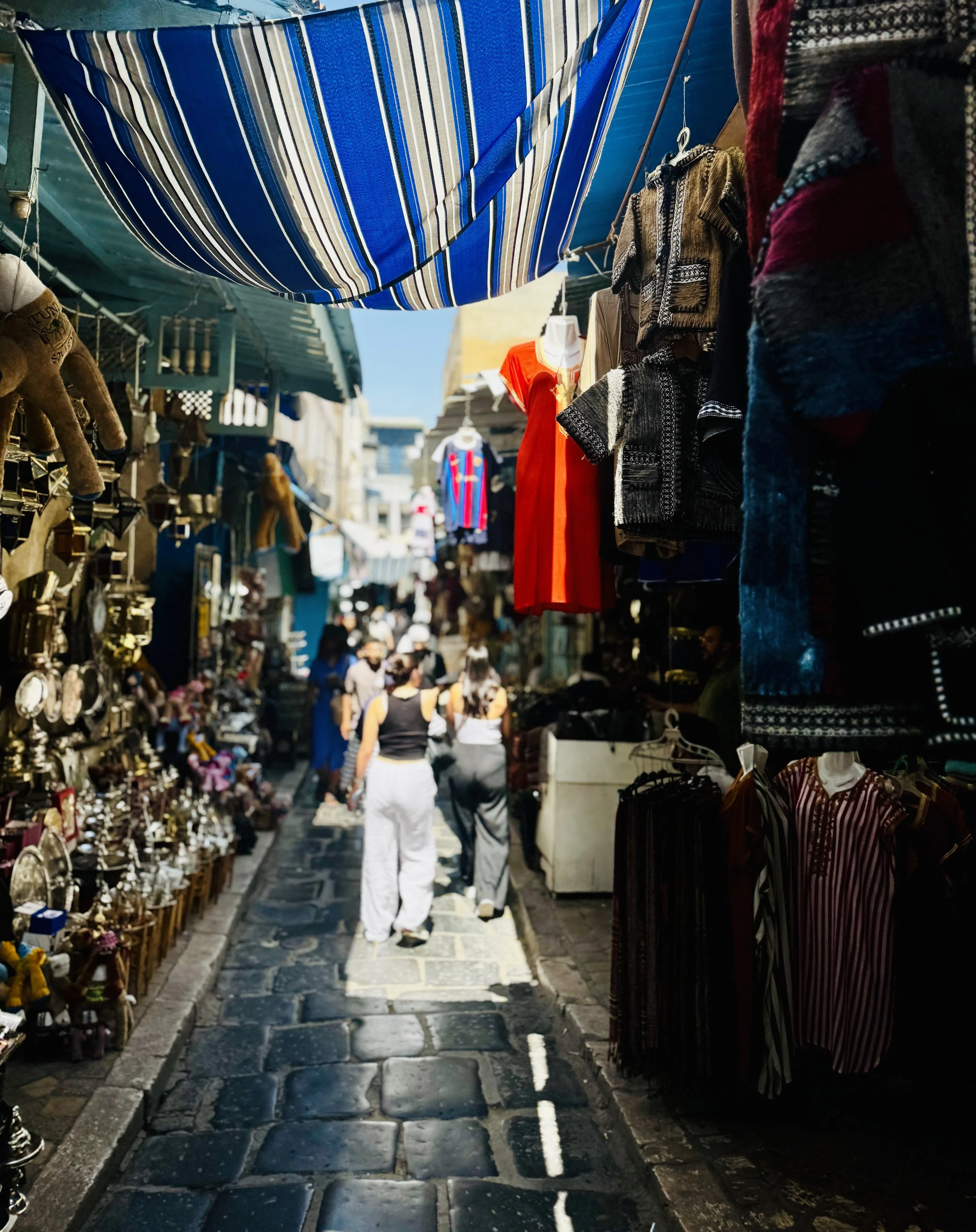 A bustling marketplace street with various stalls selling clothes and souvenirs, with people shopping and walking under colorful awnings on a sunny day.
