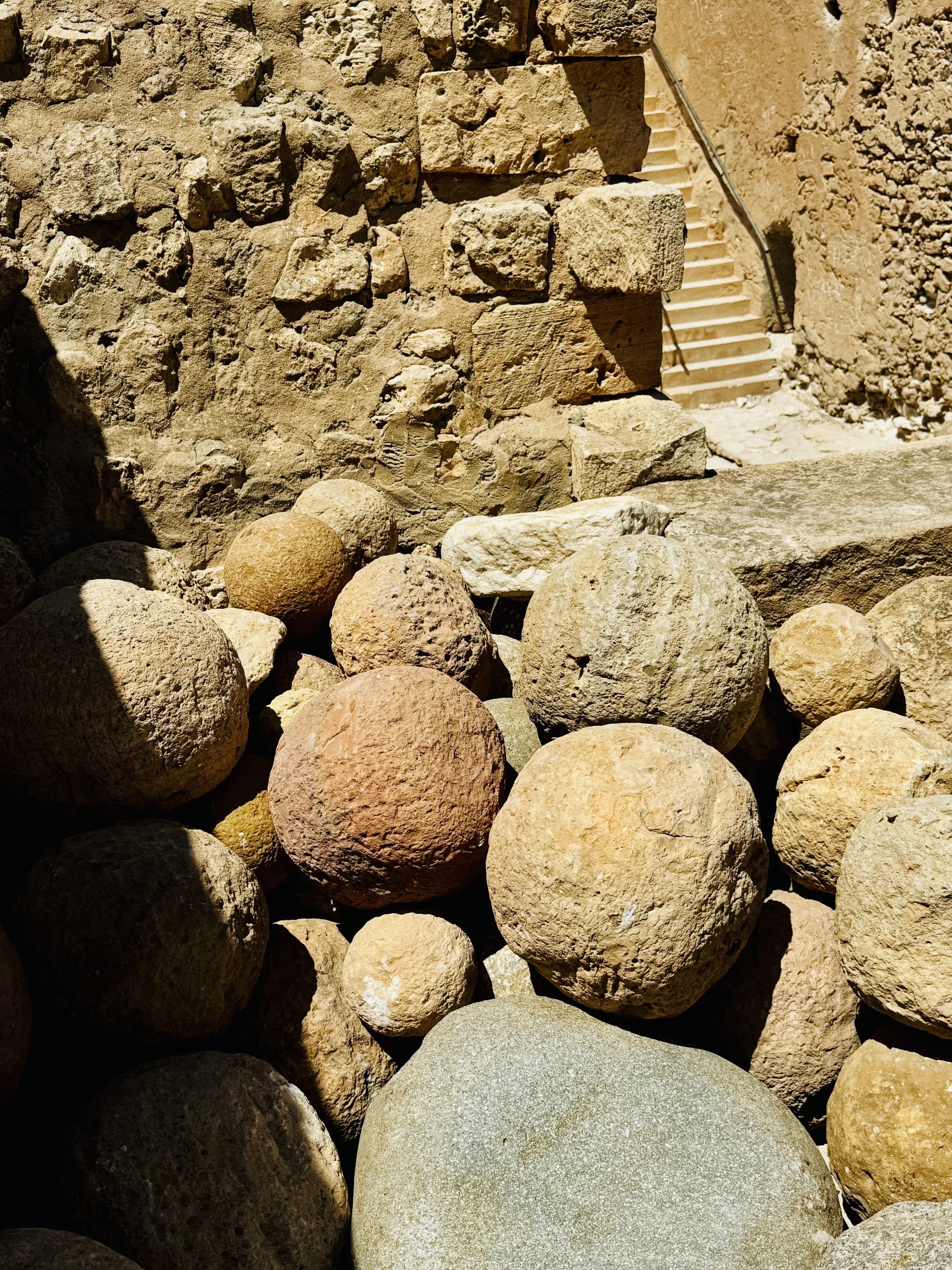 A collection of various-sized rocks, with some having rough textures and others smooth, arranged outdoors near a stone wall and staircase.