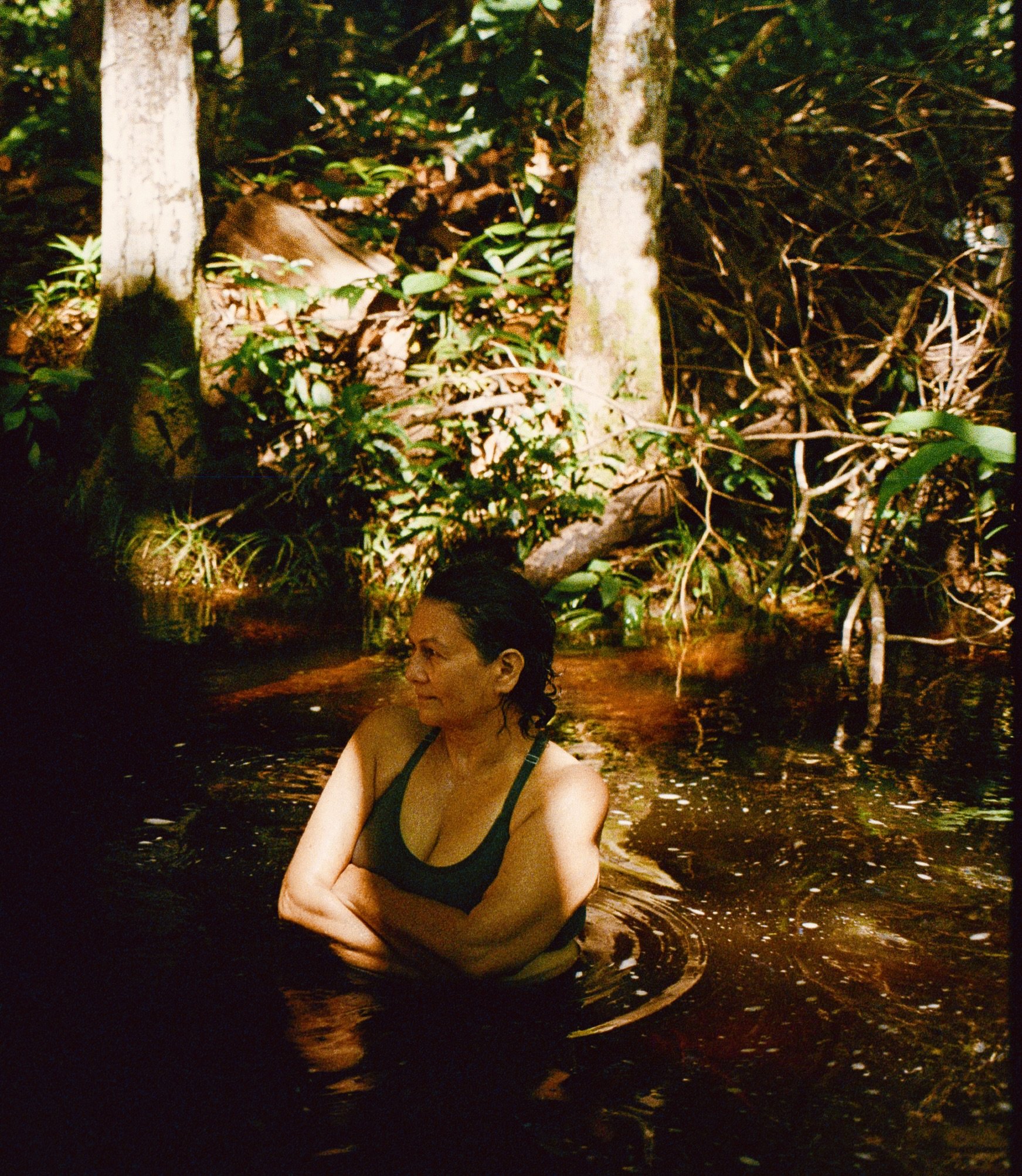 A woman with dark, wet hair and a black tank top sits in a body of water, surrounded by trees and vegetation, illuminated by artificial light at night.
