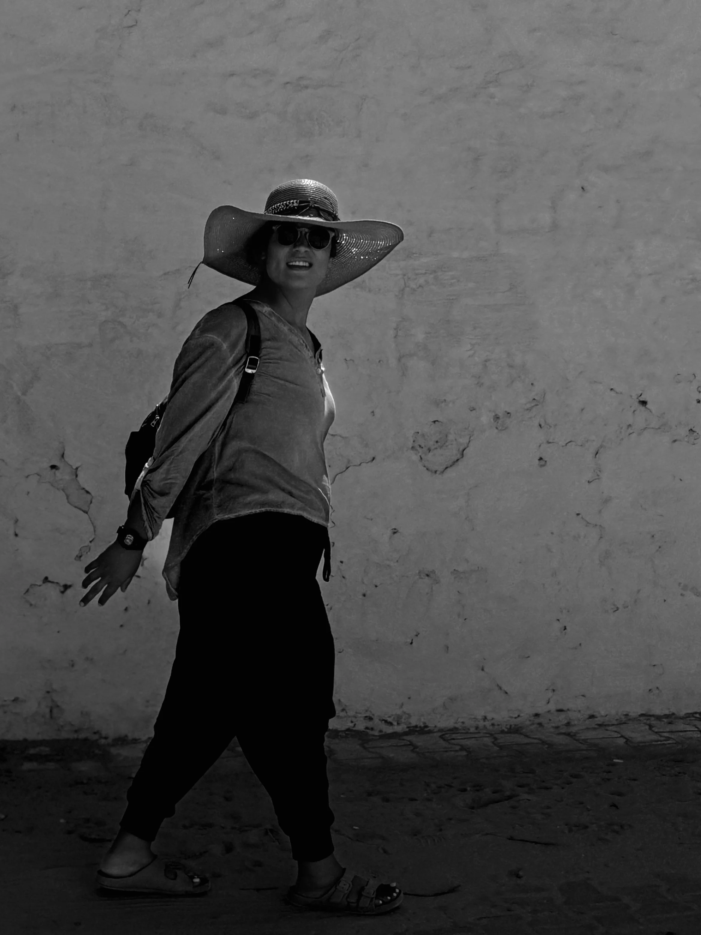 A woman wearing a large sunhat, sunglasses, a backpack, a long-sleeve shirt, and sandals, walking on a sandy surface with a plain textured wall in the background.