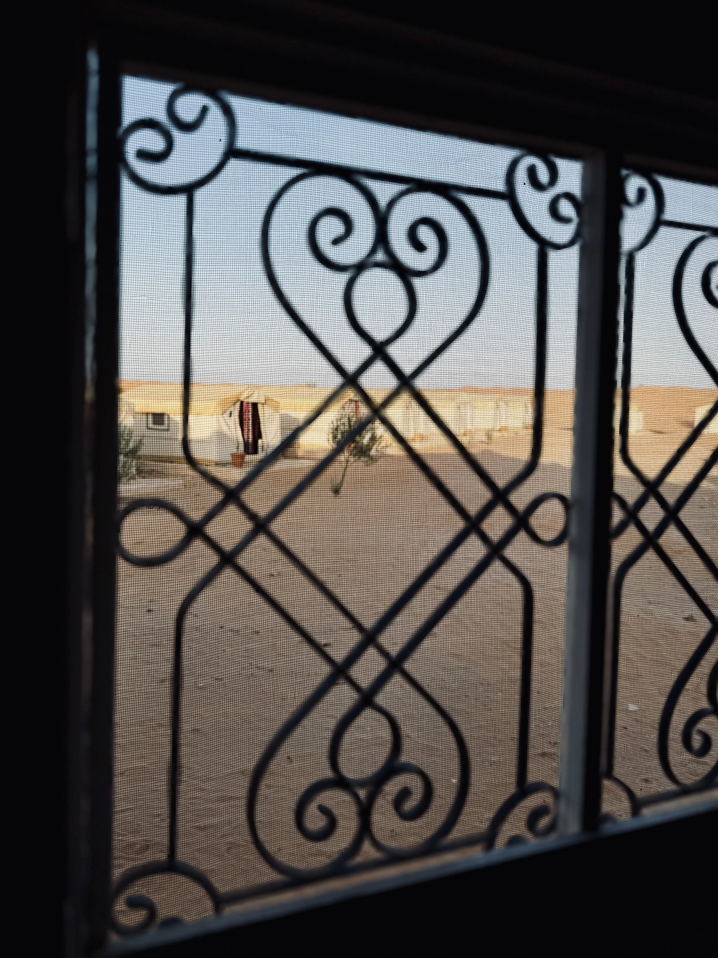 View through a decorative metal window grille showing a residential area with a house, clothes hanging on a line, and some trees in the distance.