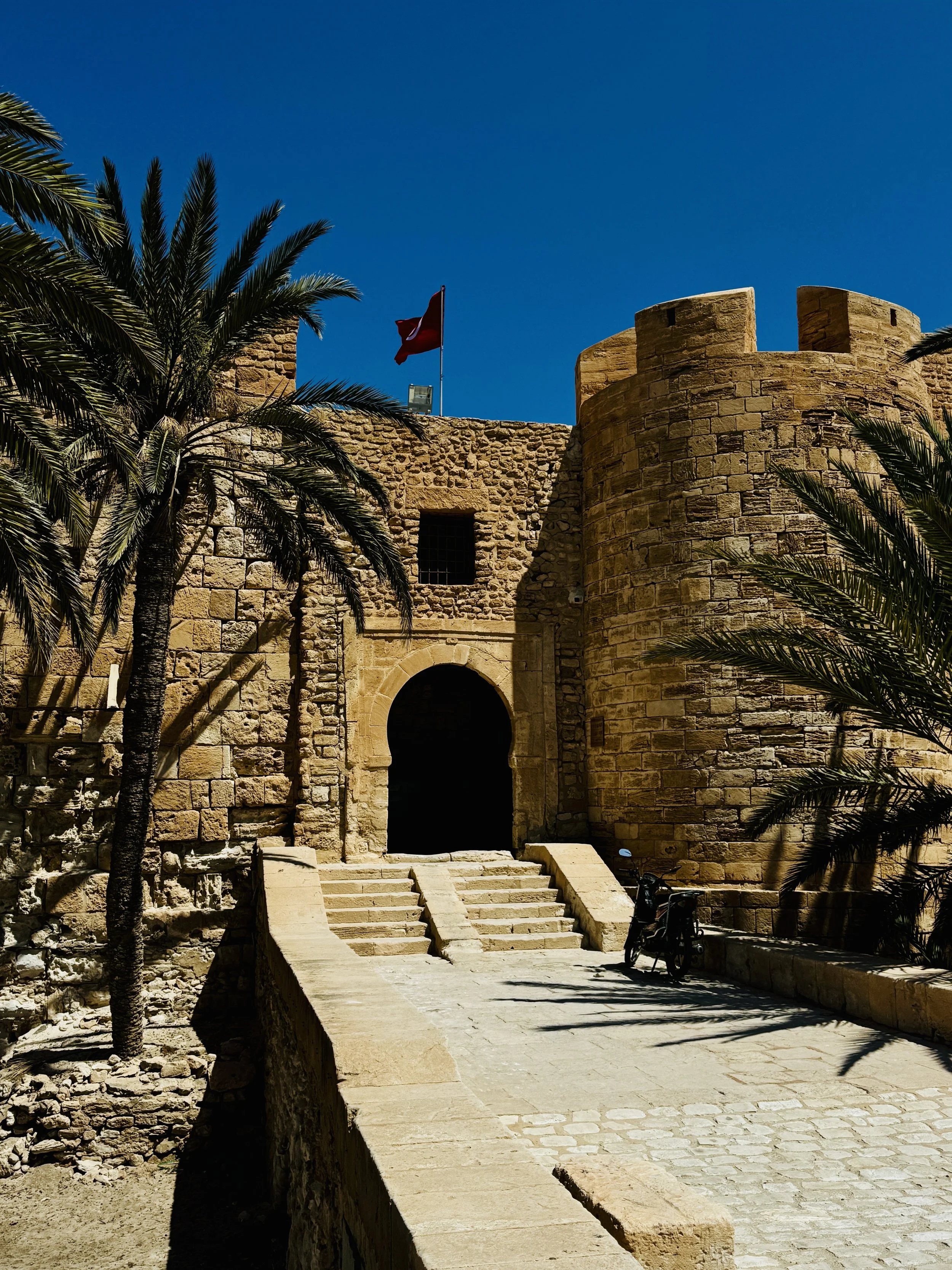 A historic stone fortress with a flag atop, palm trees, stone stairs, and an arched entrance against a clear blue sky.