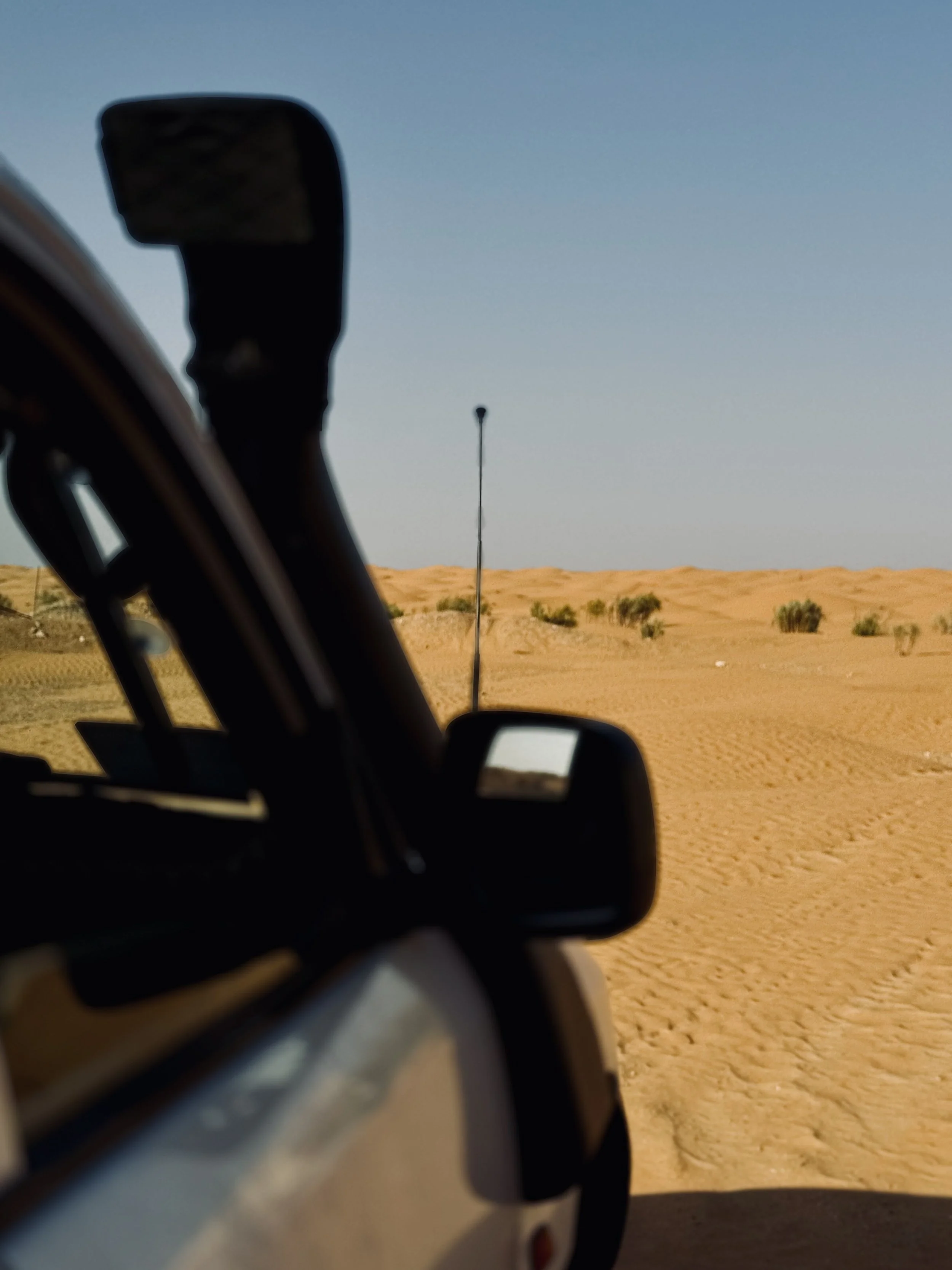 View of desert landscape with sand dunes and sparse vegetation, seen through the side mirror of a vehicle, under a clear blue sky.
