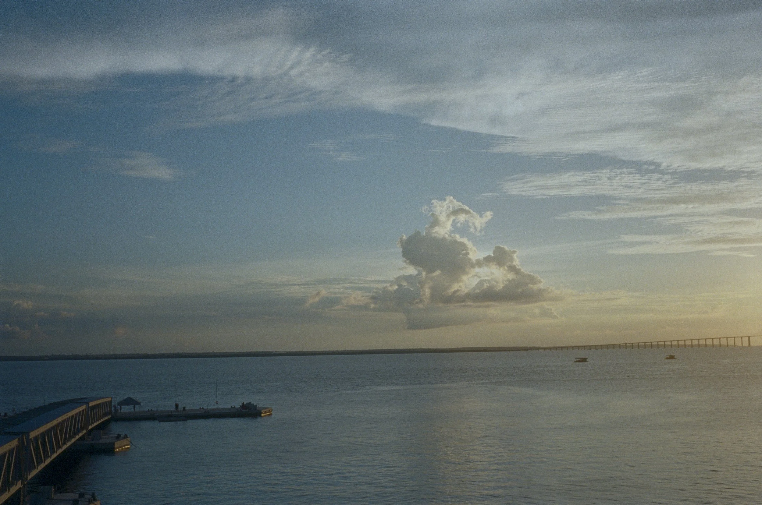 Sunset over a body of water with a pier on the left side and a bridge in the distance. Cloudy sky with scattered clouds and some sunlight.