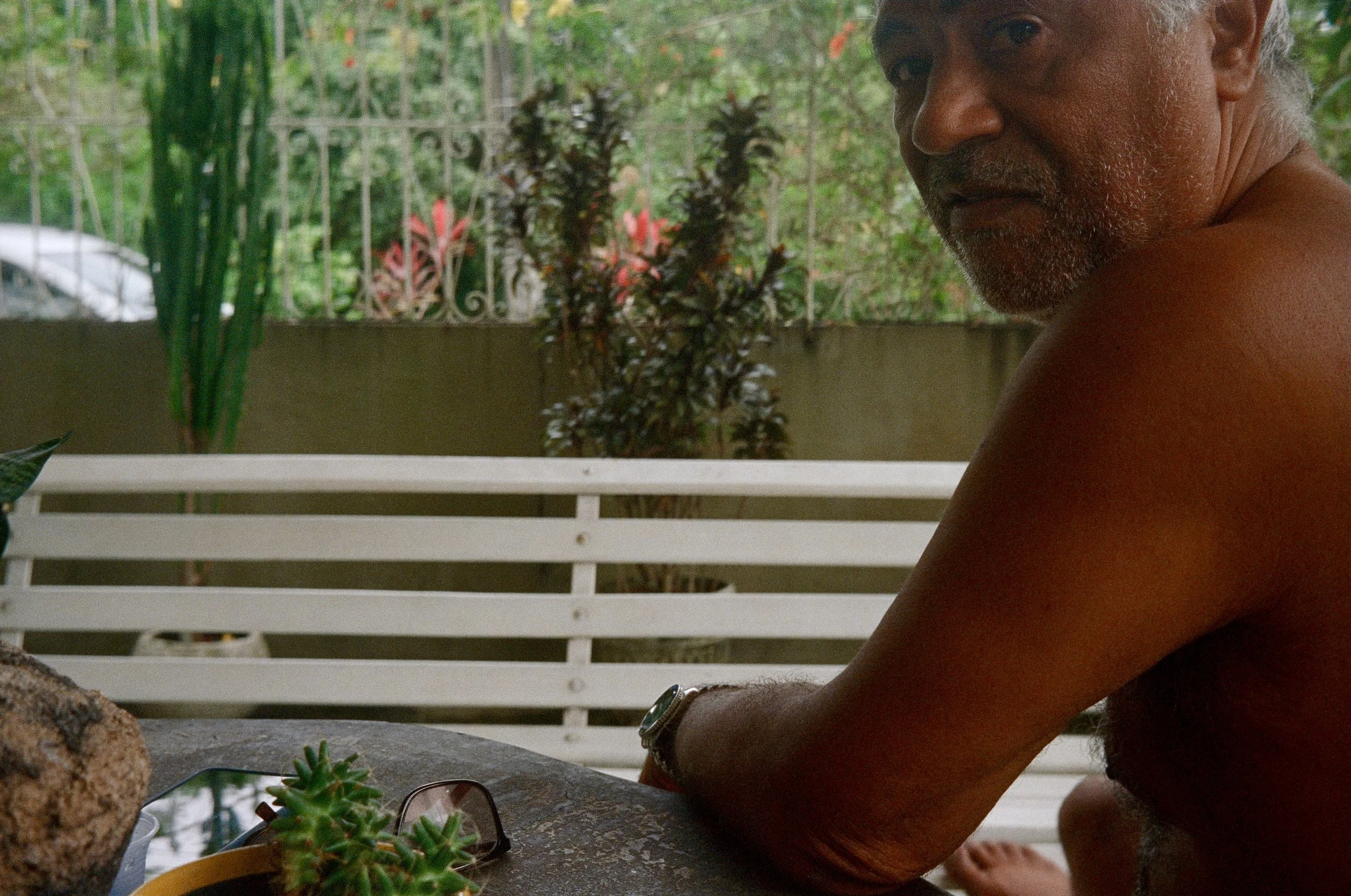A man with gray hair and a beard, shirtless, sitting outside near a white railing and green plants, looking sideways towards the camera.