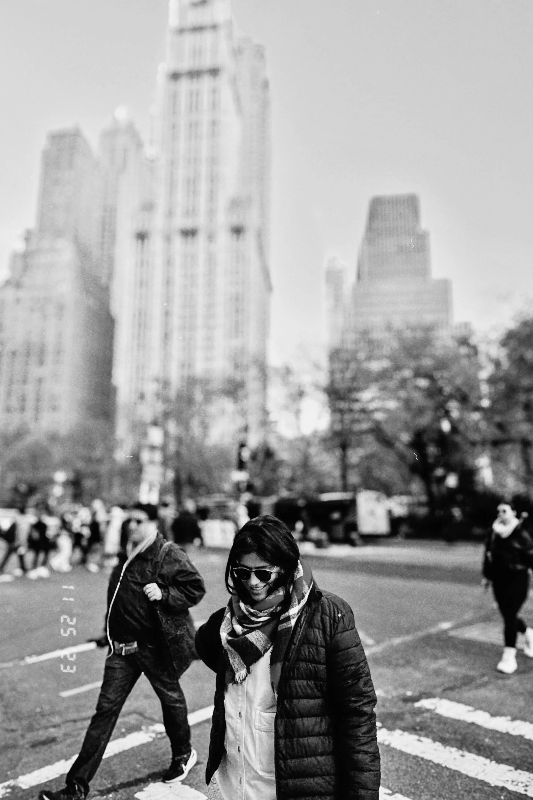 A group of people crossing a street at a crosswalk in an urban area during the daytime. In the background, skyscrapers are visible, and the weather appears clear.