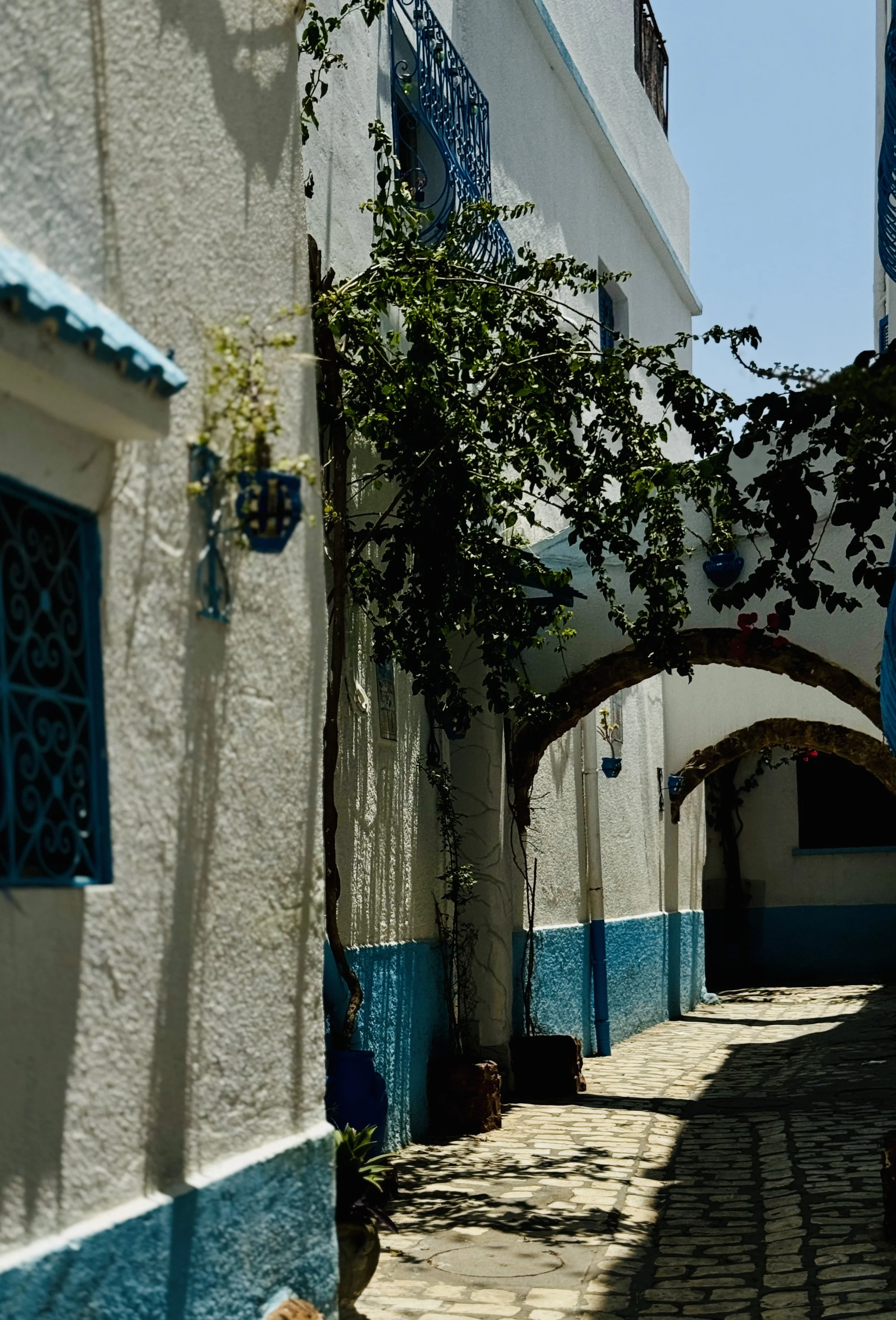 A narrow cobblestone street with white buildings featuring blue accents, arches, and decorative balconies, with shadows cast by trees and structures.