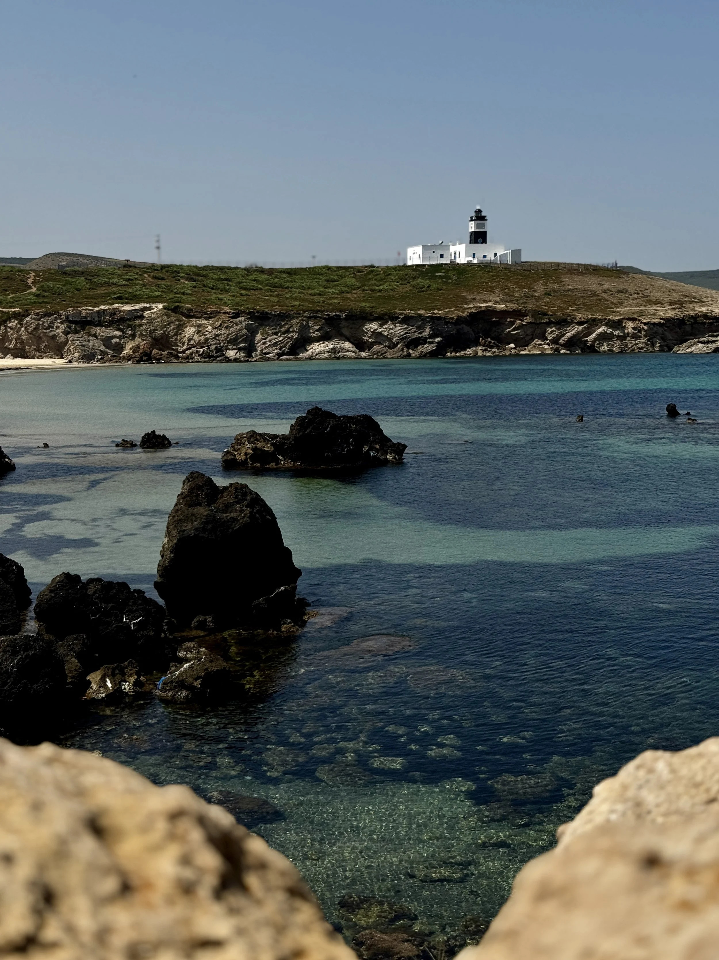A lighthouse on a hill overlooking a rocky beach with clear, turquoise water.