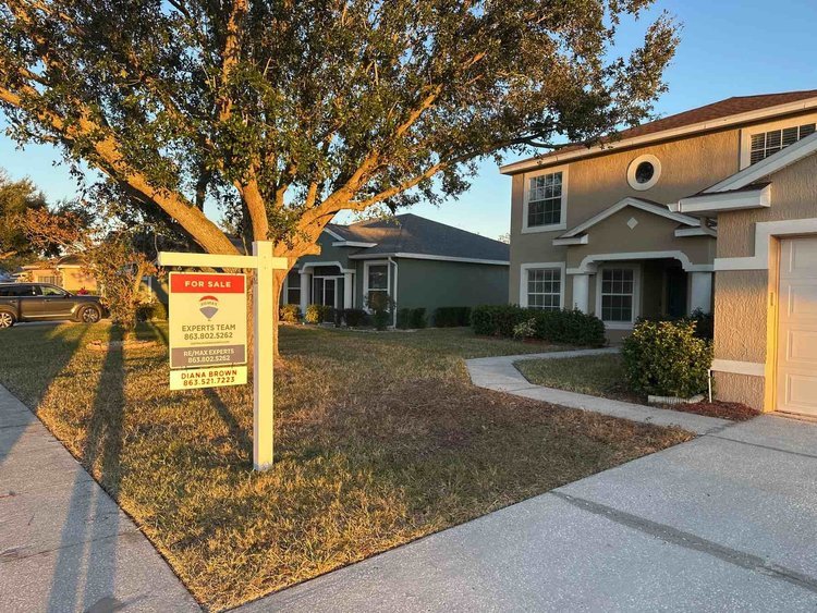 A suburban house with a yard and a for sale sign post in the front yard, with sunlight casting long shadows, a sidewalk, driveway, and parked car visible.