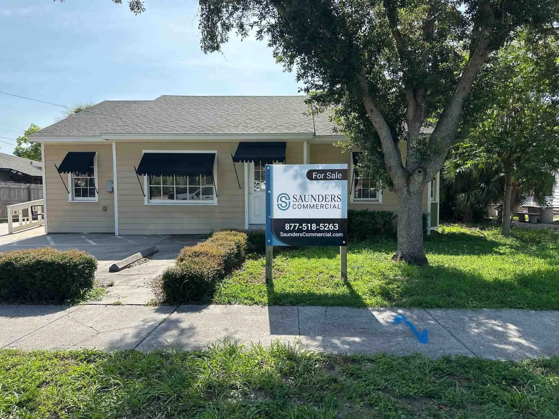 A single-story house with beige siding, black awnings over the windows, and a for sale sign in front that reads Saunders Commercial, with a phone number and website. There are trees and bushes around the house, and a sidewalk with a blue arrow pointing towards the house.