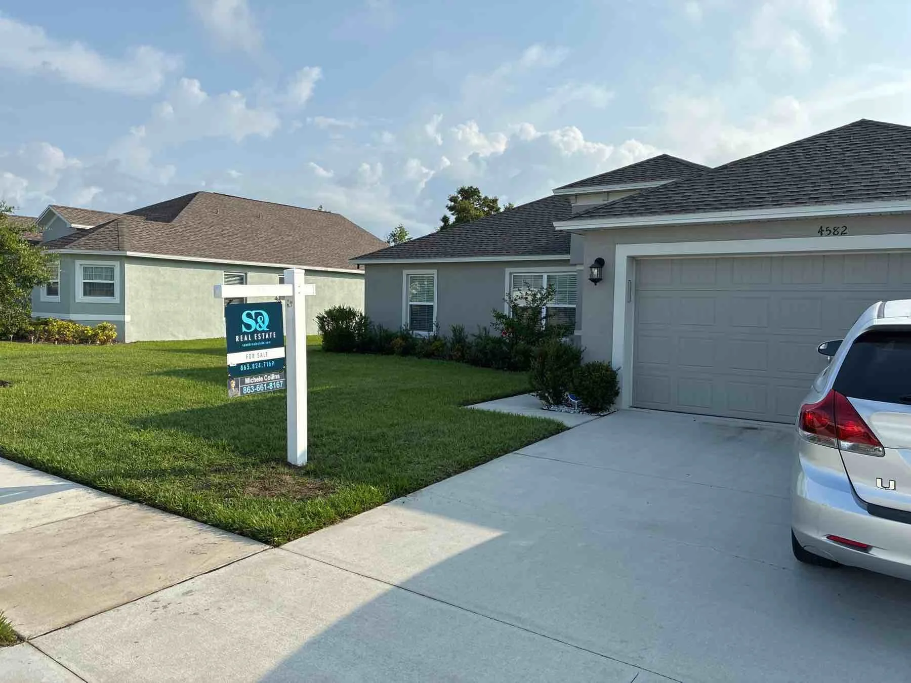 A house with a light gray exterior, a brown shingled roof, a garage door, a white car parked in the driveway, a lawn with green grass, and a real estate sign that reads 'S&Q Real Estate for Sale' with contact information. The sky is partly cloudy.