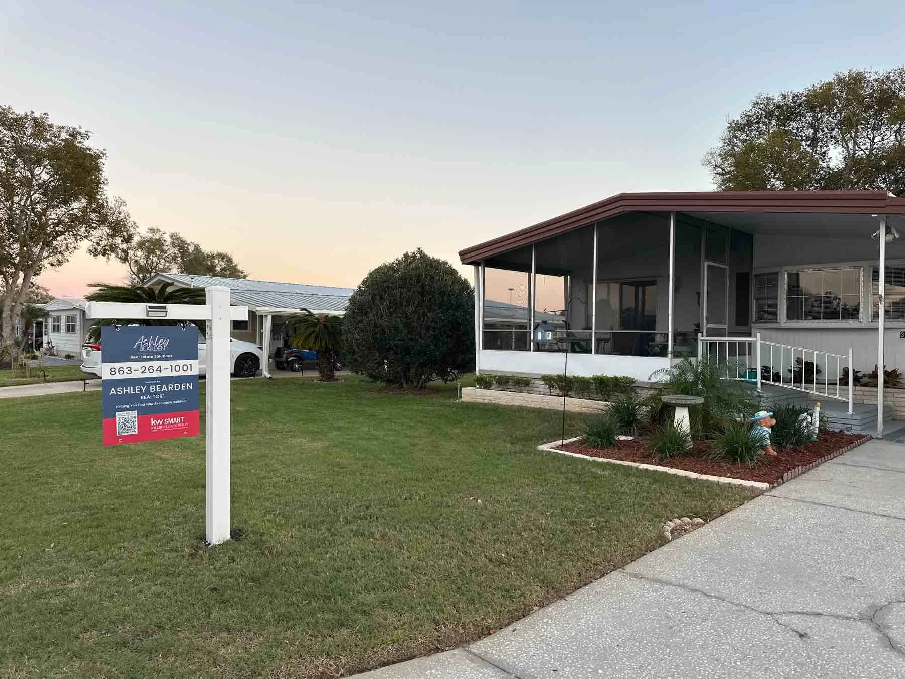 A residential house with a porch, surrounded by a landscaped lawn with small plants and a statue of a gnome. A sign post in the front yard advertises real estate services for Ashley Bearden, a realtor.