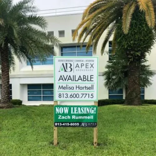 A sign in front of a building advertising apartments for lease. The top part of the sign shows the logo of APEX it also says "Available" with the name Melisa Hartsell and a phone number. The bottom part of the sign says "Now Leasing!" with Zach Rummell's name and contact number.