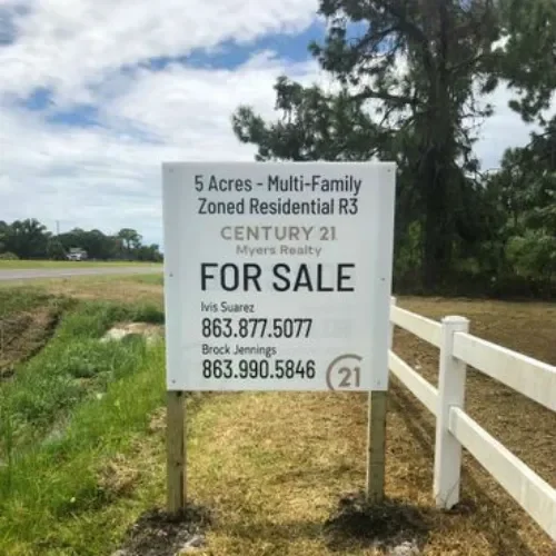 Sign advertising 5 acres of land for sale, zoning R3, contact numbers for realtors, with trees and grassy field in the background.