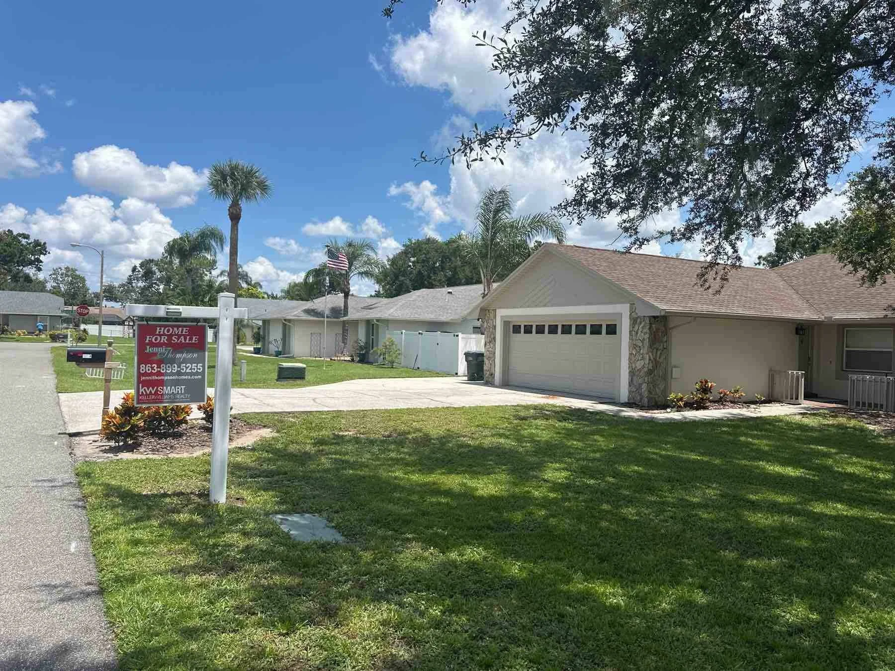 Single-story house with a garage, a lawn, and a 'Home For Sale' sign post in the front yard, under a partly cloudy sky with trees and palm trees nearby.
