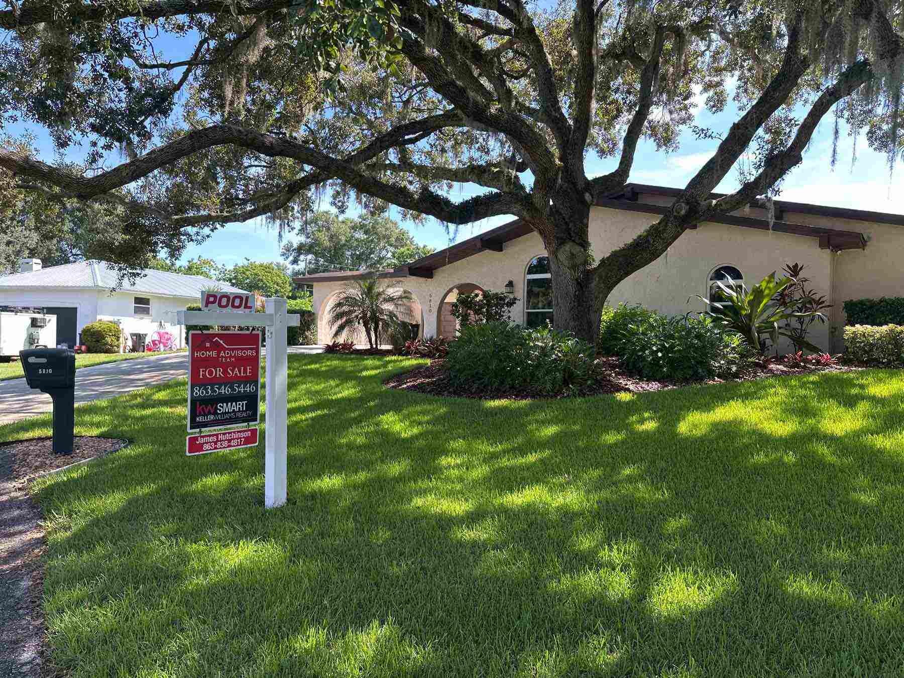 A house with a landscaped front yard, large tree, and a 'For Sale' sign post in the lawn. The sign indicates the property is listed by Keller Williams Realty.