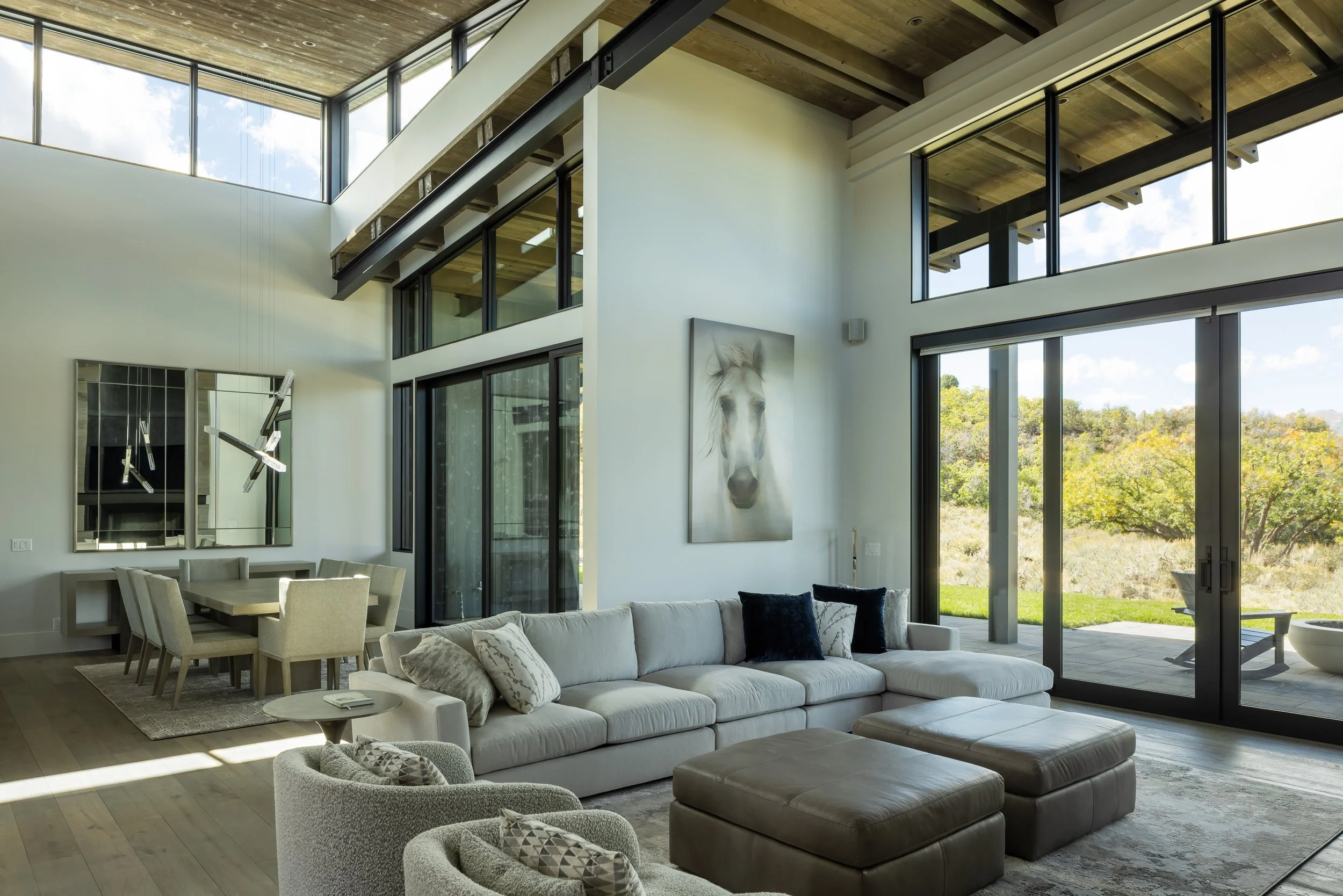 Modern living room with large windows, white sectional sofa, tan ottomans, artwork of a horse, dining area with beige chairs, and a view of trees outside.