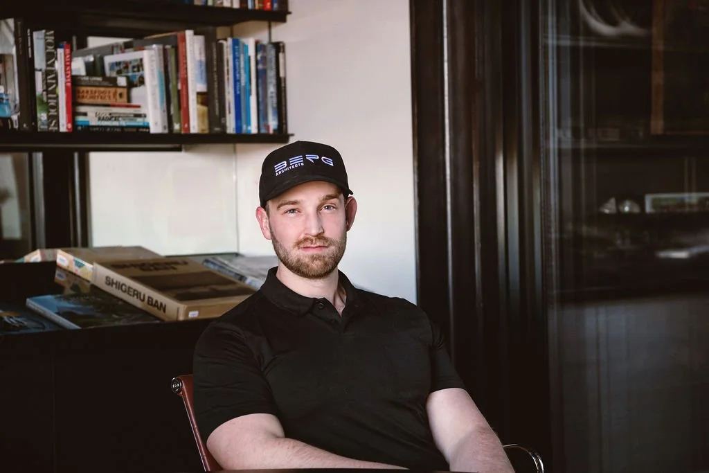A man with a beard and mustache wearing a black polo shirt and a black cap sitting in a room with bookshelves and a black cabinet.