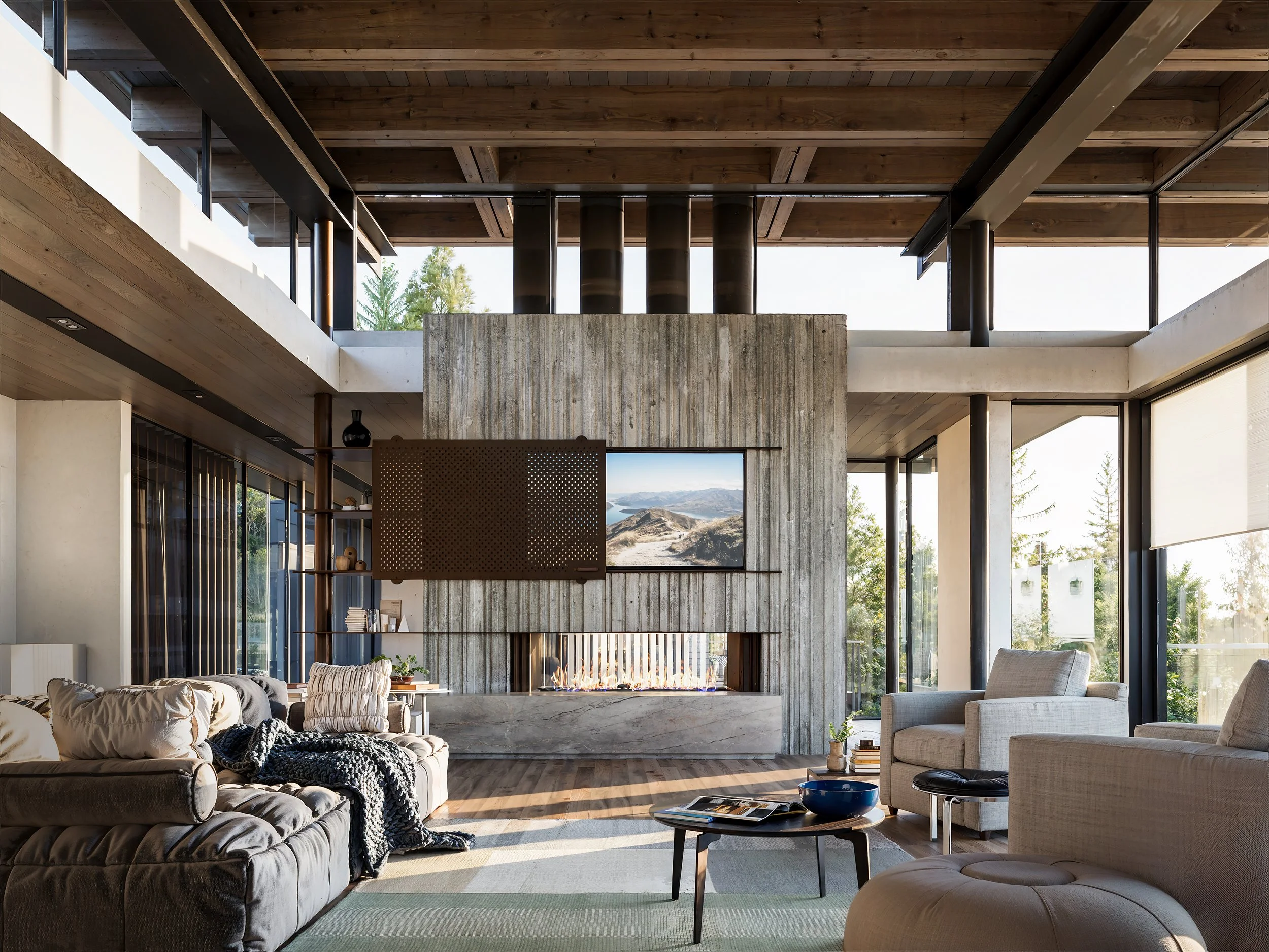 Modern living room with concrete fireplace, flat screen TV, beige sofas, armchair, coffee table, and large windows showing trees outside.