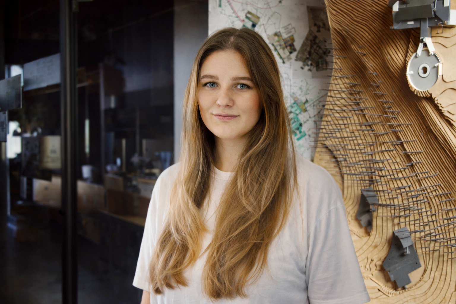 A young woman with long, wavy blonde hair and blue eyes standing indoors in front of a wooden art installation with metal spikes and a map on the wall behind her.