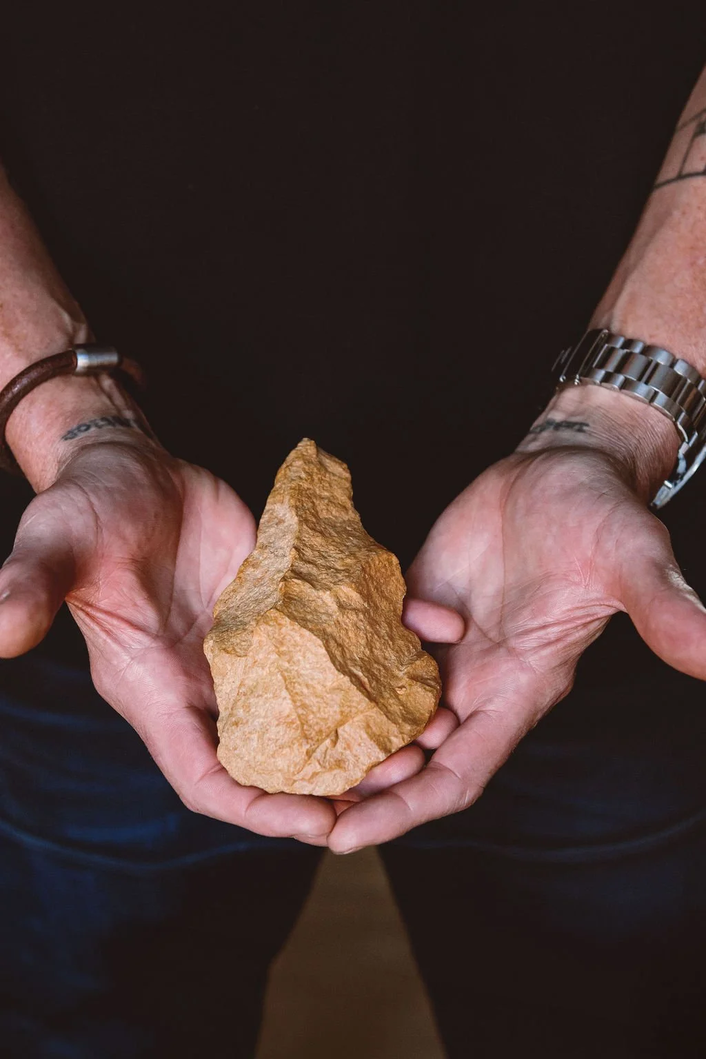 Person holding a large, brown rock with both hands, wearing a watch and bracelet on their wrists.