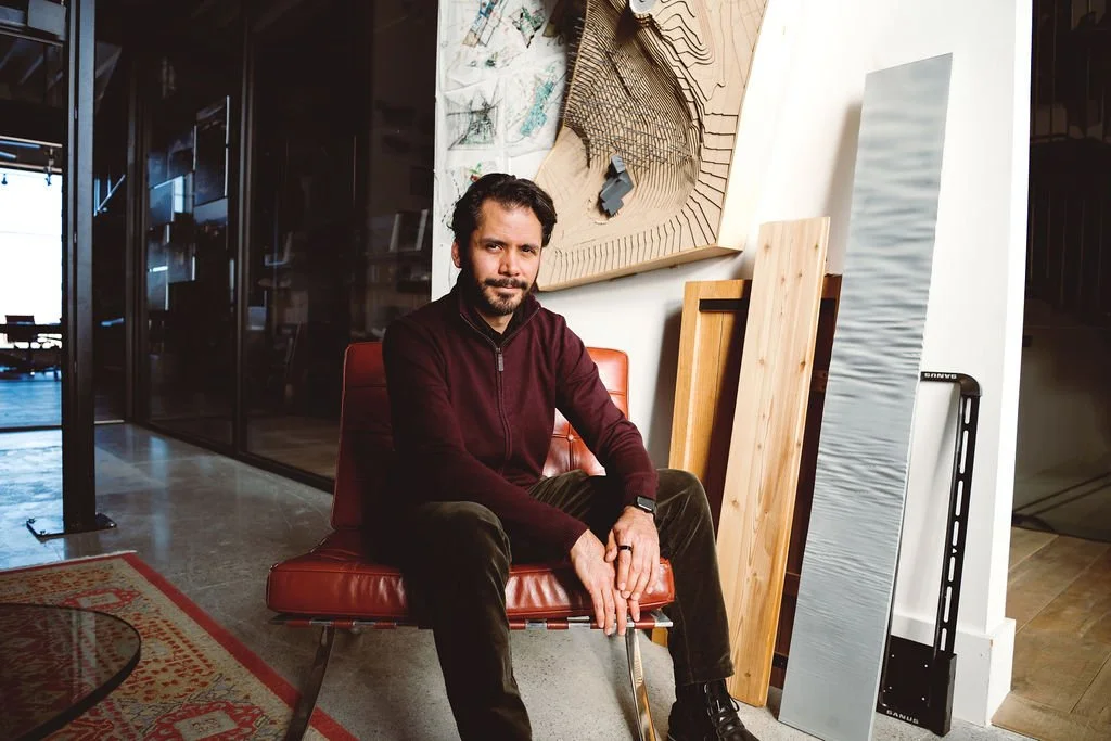 A man with dark hair, beard, and wearing a maroon sweater and black pants, sitting on a red chair in a modern art studio or office space with wood and architectural models around him.