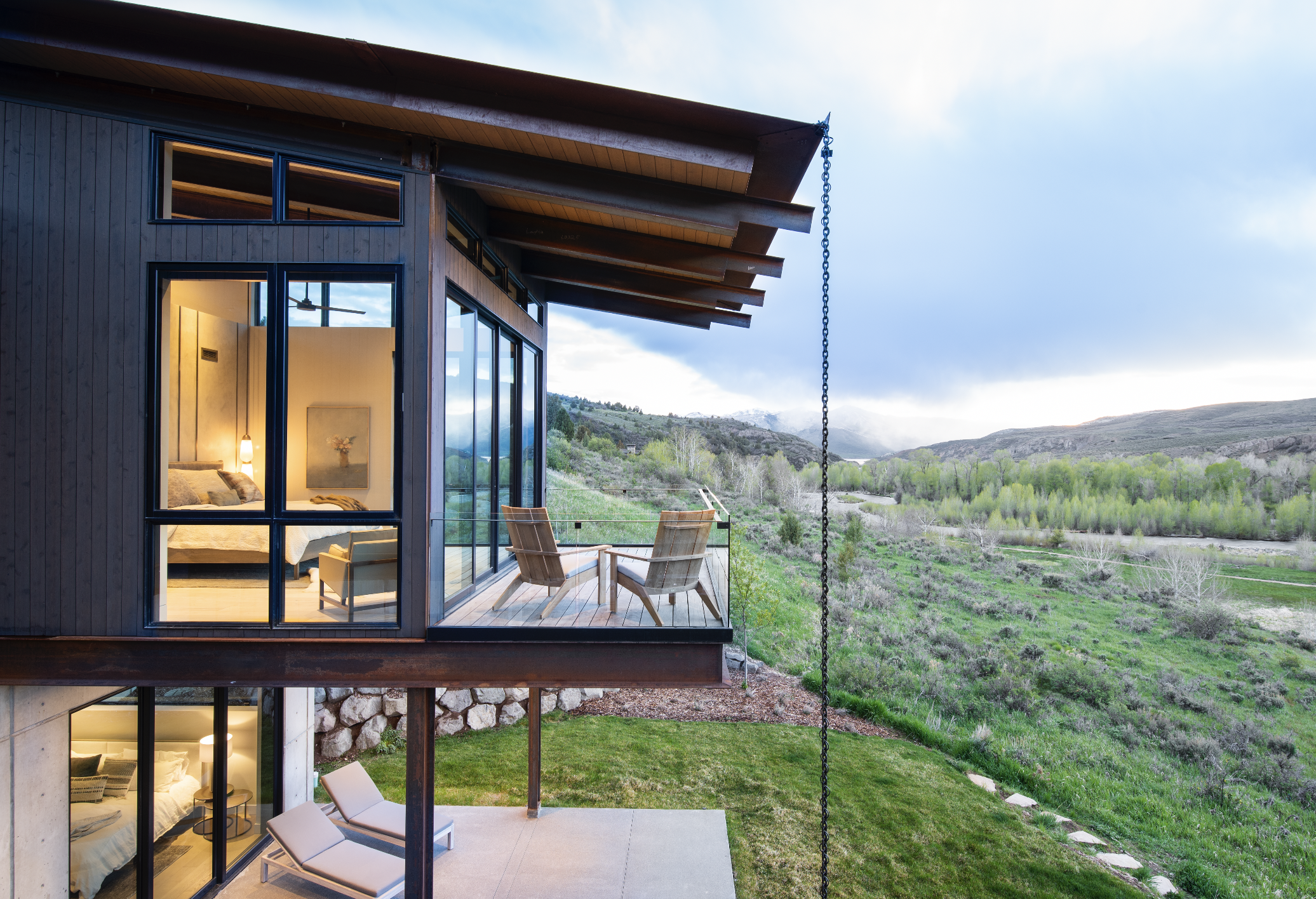 Modern house with large glass windows and a balcony overlooking a green landscape with hills and trees, with interior lighting visible inside.