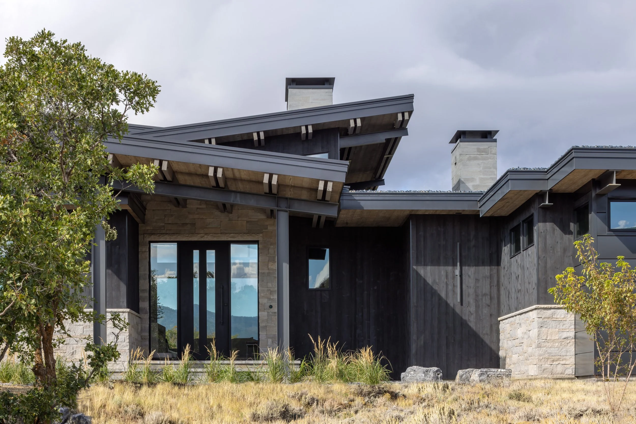 Modern house with geometric rooflines, large glass doors, dark wood siding, stone accents, and a landscaped yard with grasses and trees.