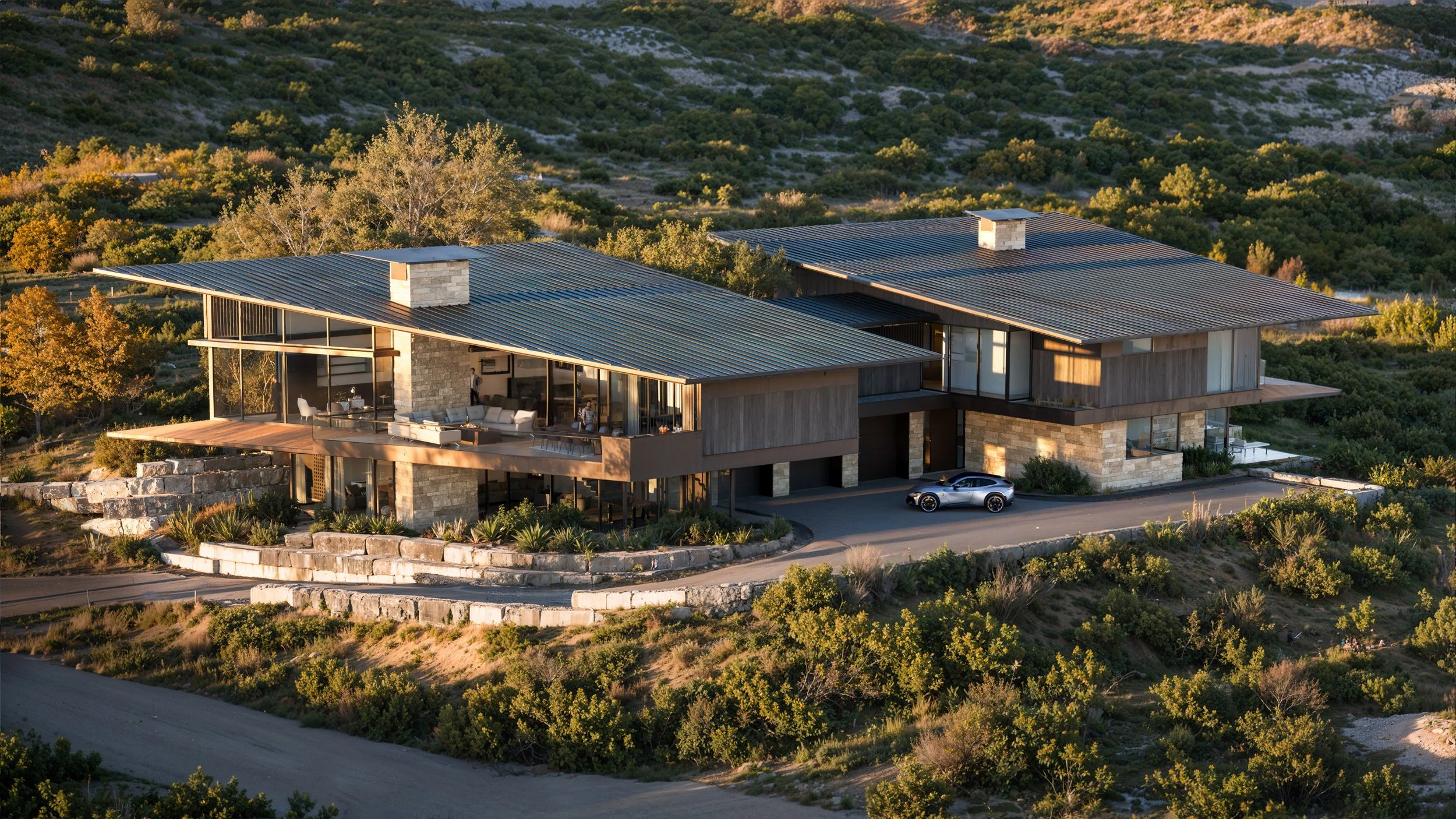 Modern house with large glass windows, stone exterior, and metal roofing, situated on a hillside with trees and shrubbery, and a car parked in front.